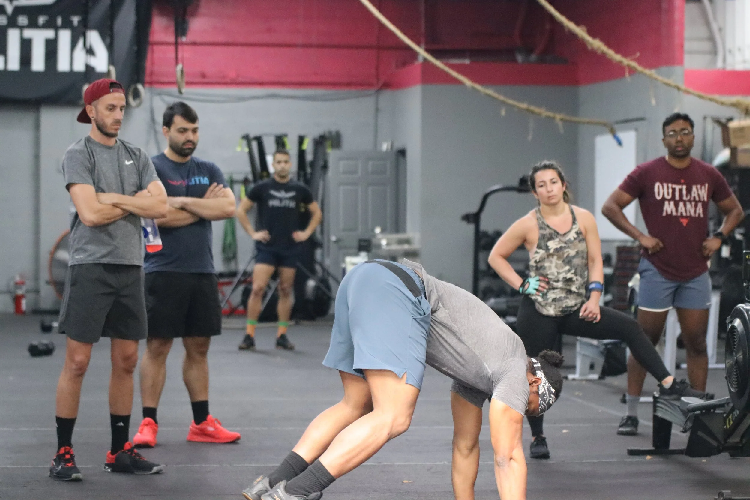 People in a gym with one person bending over during workout, surrounded by onlookers.