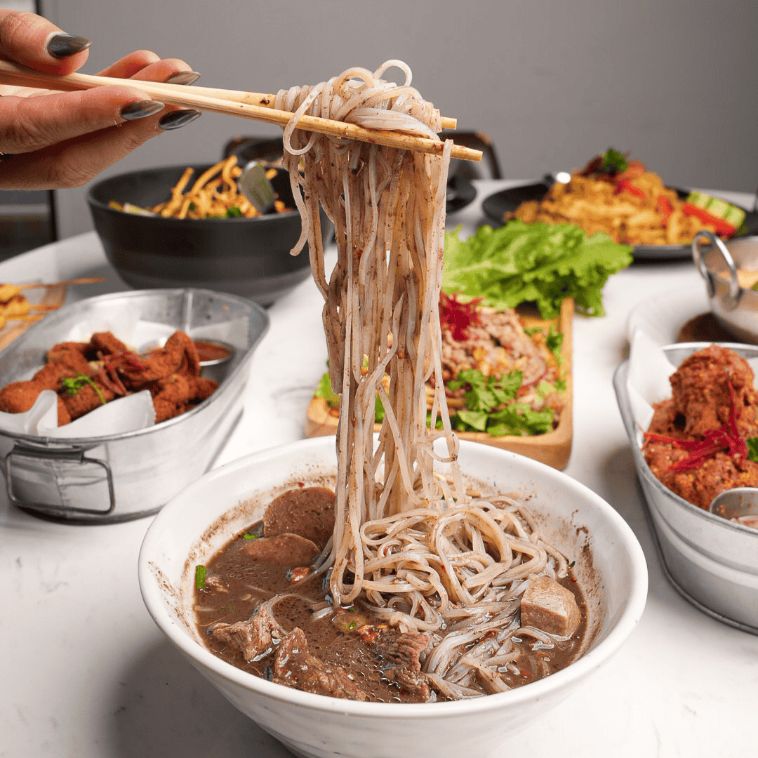 A person lifting noodles from a bowl of beef noodle soup with chopsticks, surrounded by various Asian dishes including fried chicken, lettuce, and other noodle dishes on a white table.