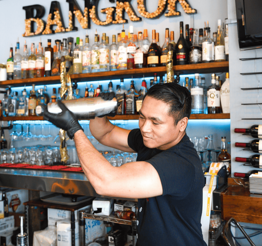 A bartender with black hair, wearing a black t-shirt and black gloves, shaking a cocktail shaker behind a bar with liquor bottles on shelves in the background.