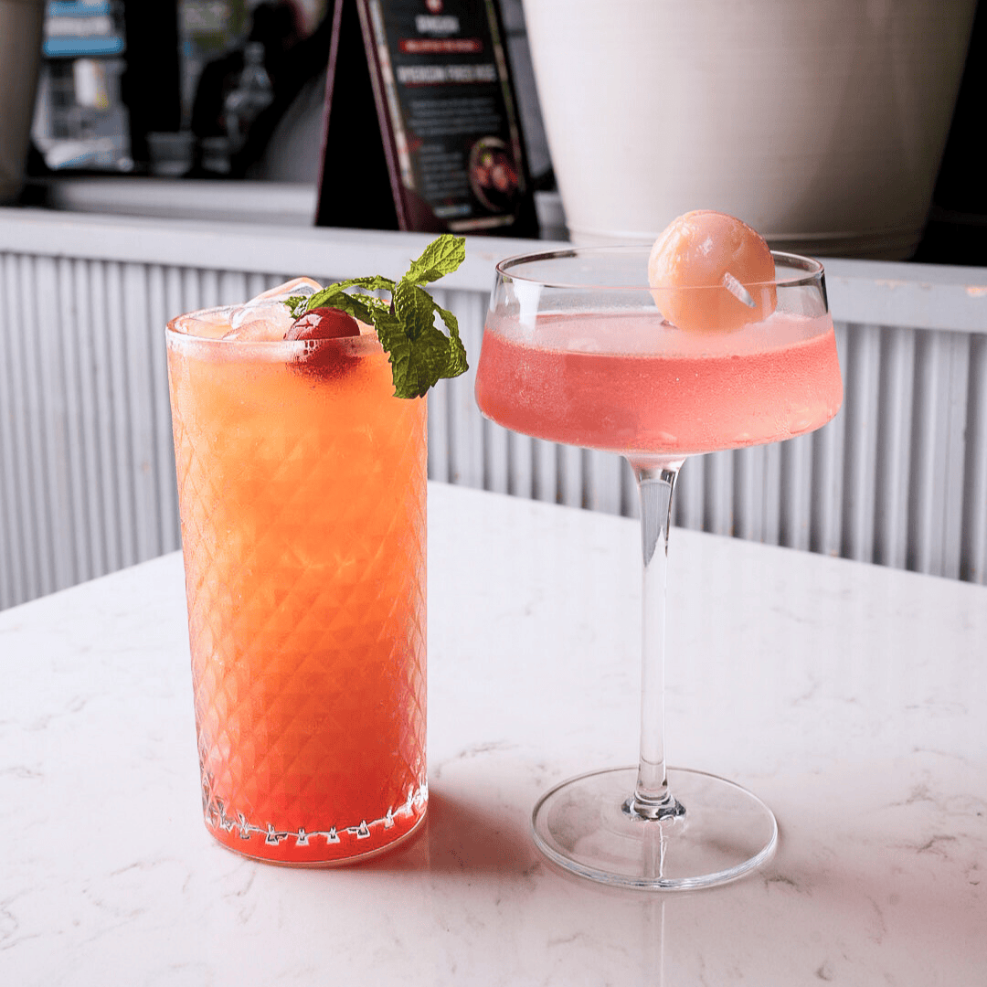 Two pink cocktails, one in a tall textured glass garnished with mint and a cherry, the other in a coupe with a cherry on top, on a white marble table.
