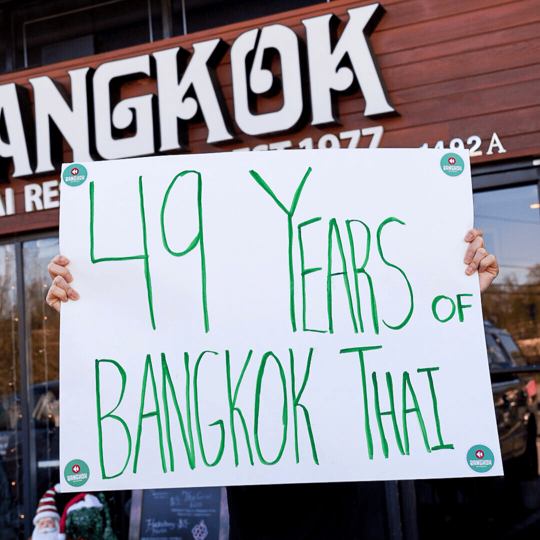 Person holding a white sign that reads '49 Years of Bangkok That' in front of what appears to be a restaurant or storefront.