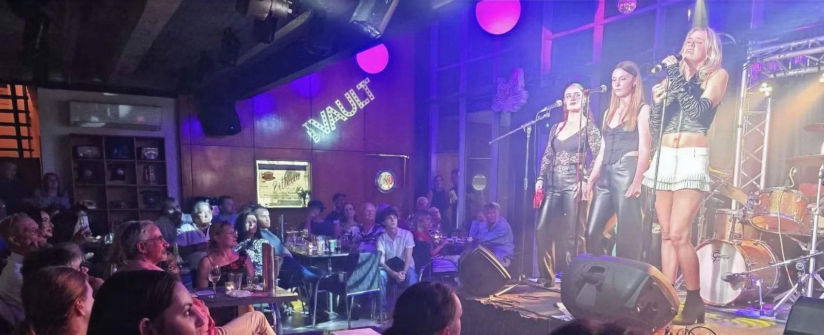 Three women singing on stage at a dimly lit performance venue, with an audience seated at tables watching them.