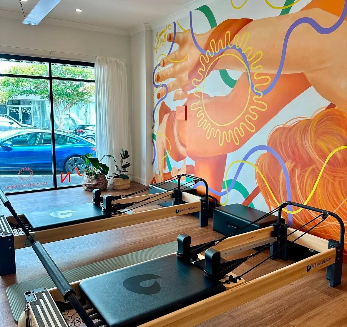 A Pilates studio with two reformer machines and a colorful abstract mural on the wall, featuring a large hand with orange, pink, and yellow tones, and squiggly lines in purple, green, and yellow. Potted plants are near the glass door, and cars are visible outside.