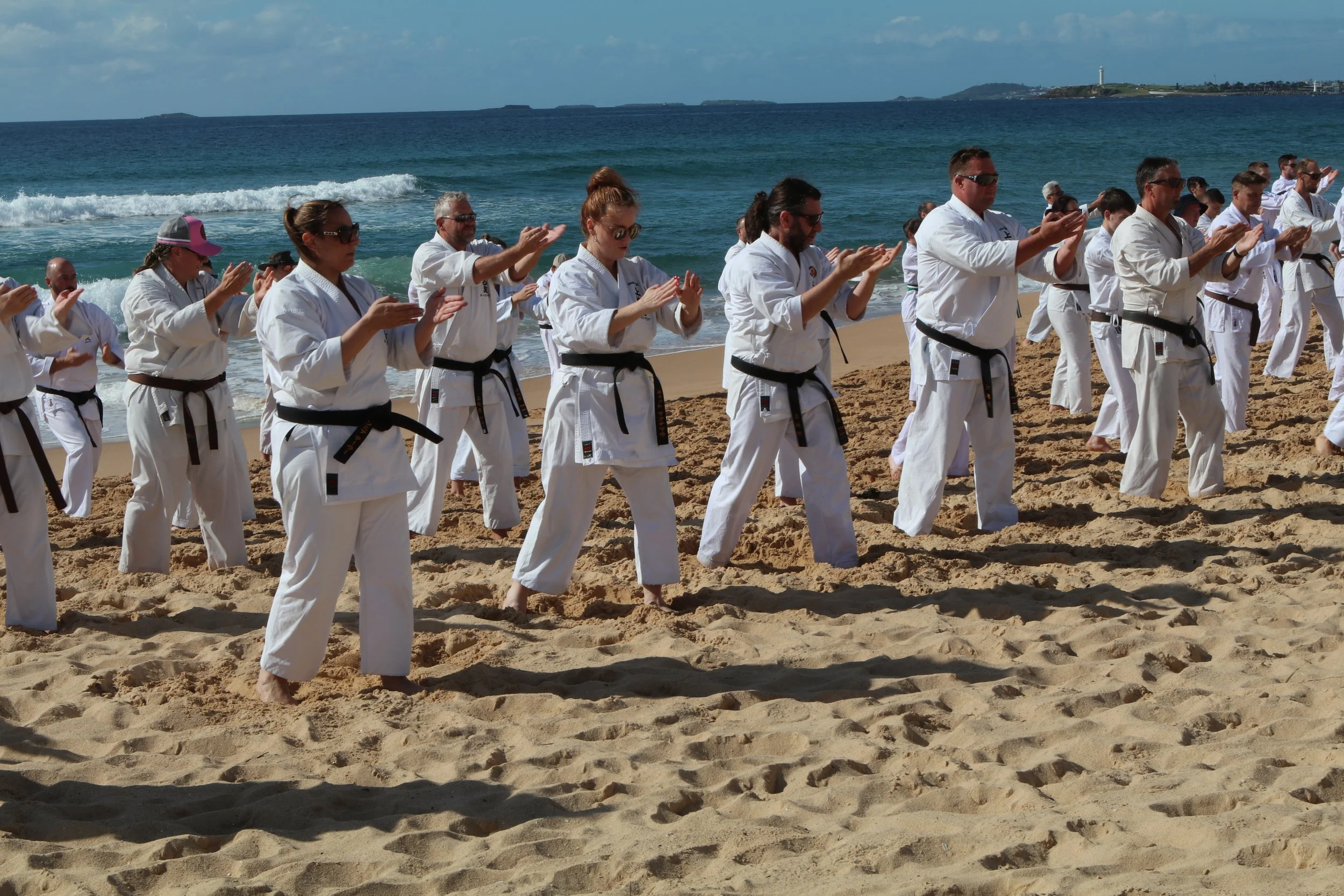 People practicing martial arts on the sandy beach during the daytime with the ocean and a lighthouse in the background.