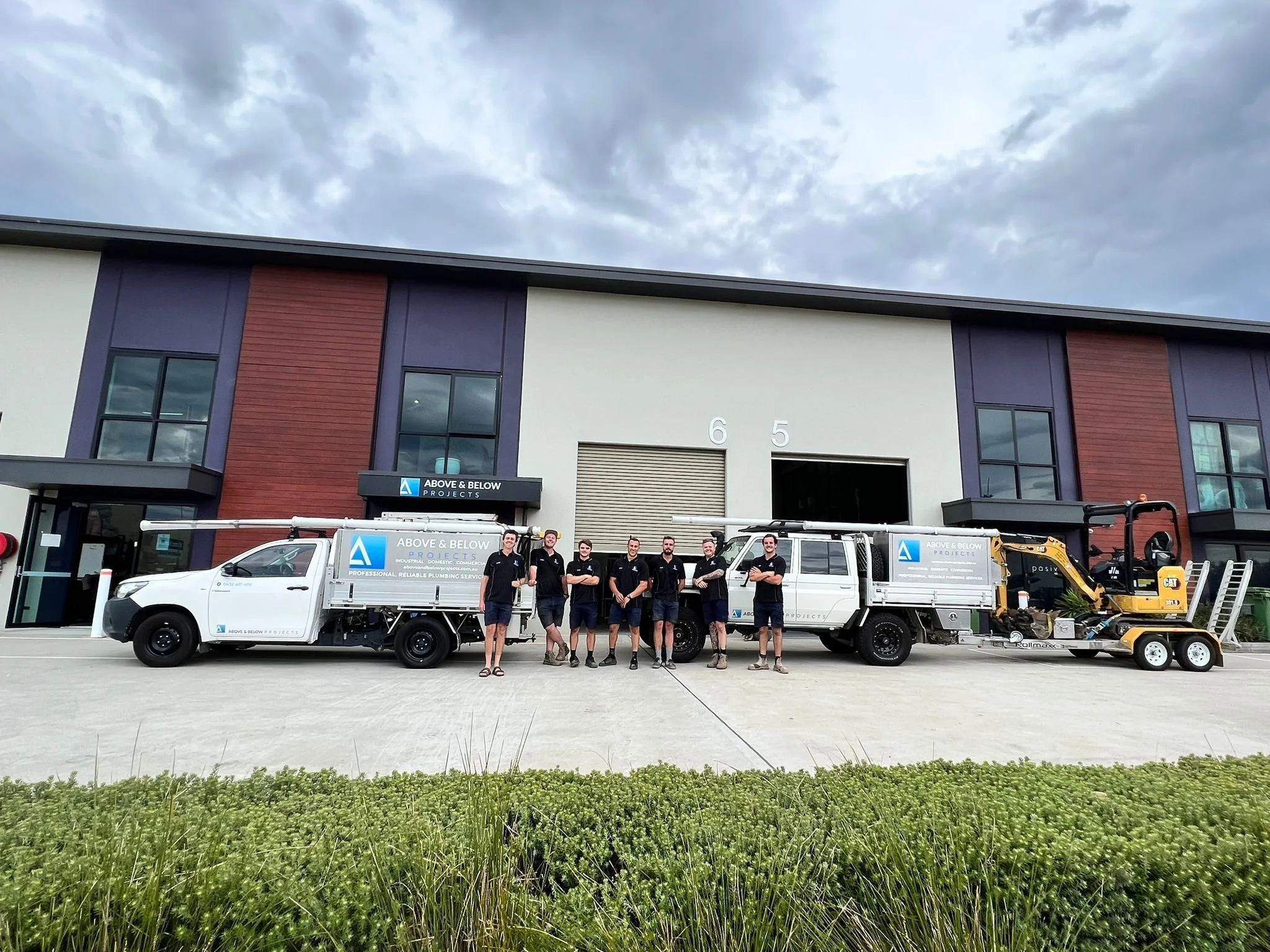 Group of workers standing in front of company trucks and building, with cloudy sky overhead.