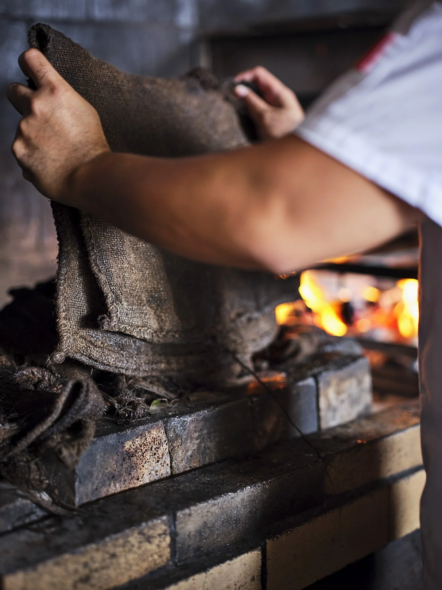 A person handling a piece of meat or food item over an open flame, with a brick or stone surface below, in a kitchen or outdoor cooking setting.