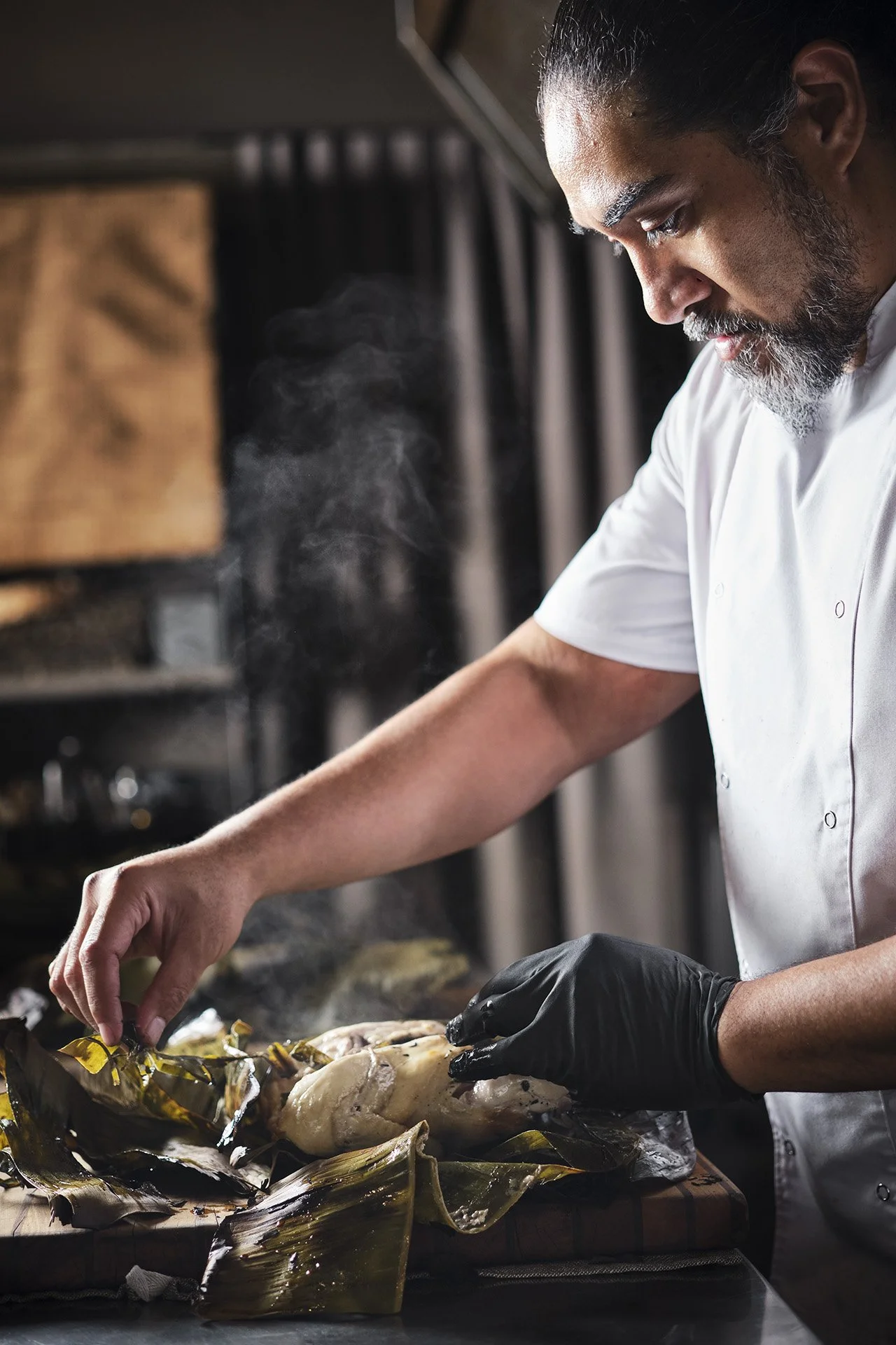 A chef in a white uniform preparing a dish with a piece of fish on a cutting board, wearing black gloves, in a professional kitchen.