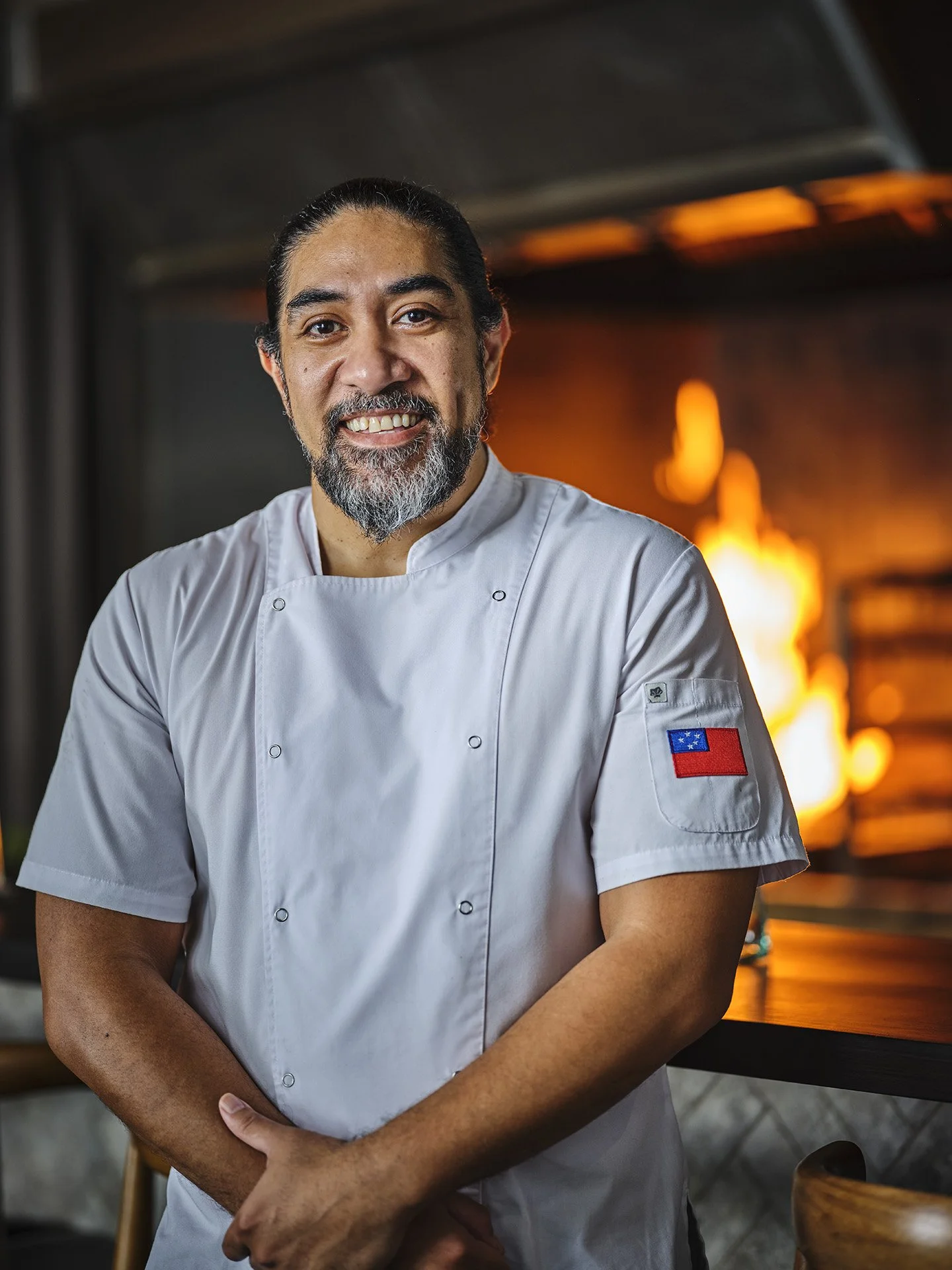 A smiling male chef with long dark hair and a beard, wearing a white chef coat with an embroidered patch of the American flag on the sleeve, standing in front of a fiery kitchen with an orange glow.