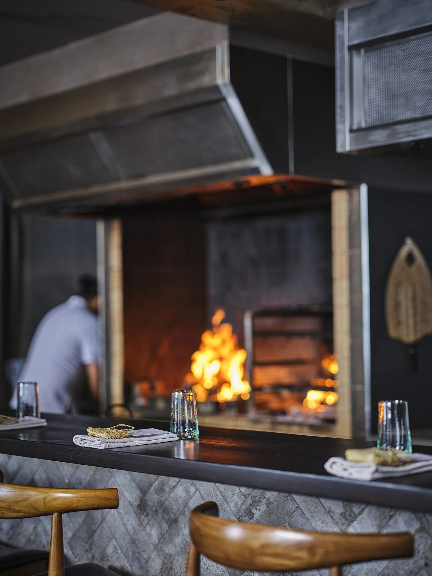 Interior of a restaurant with a wood-fired grill in the background, a man preparing food, and a counter with glasses and napkins.