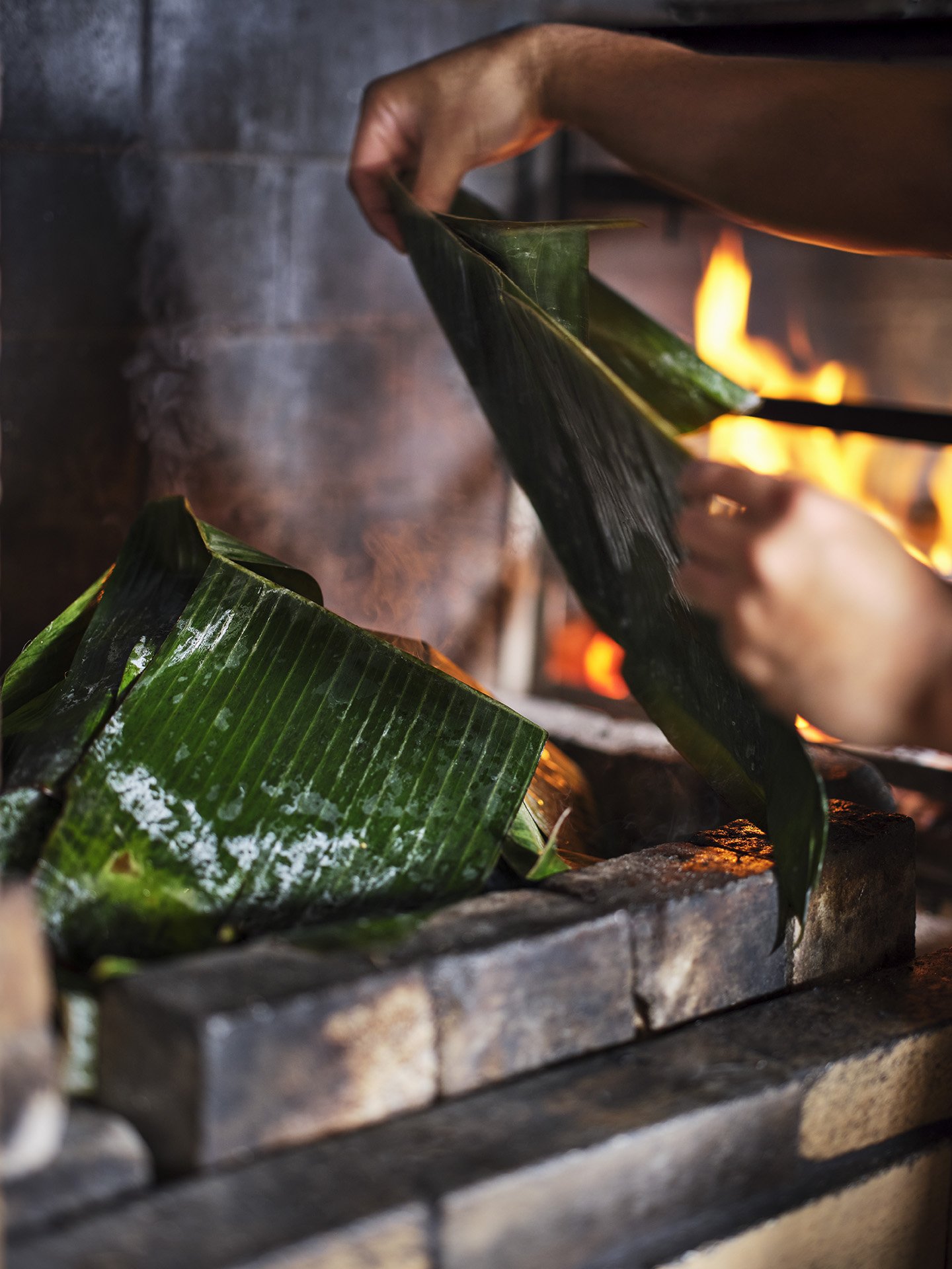 Close-up of hands roasting banana leaves over an open flame in a brick fireplace.