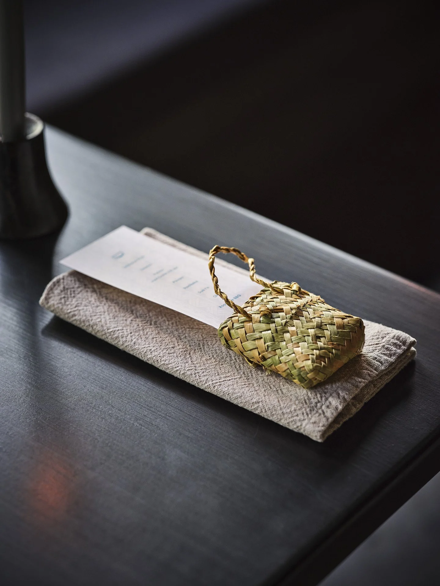A woven straw bag resting on a folded beige cloth, with an envelope or paper underneath on a dark wooden surface.