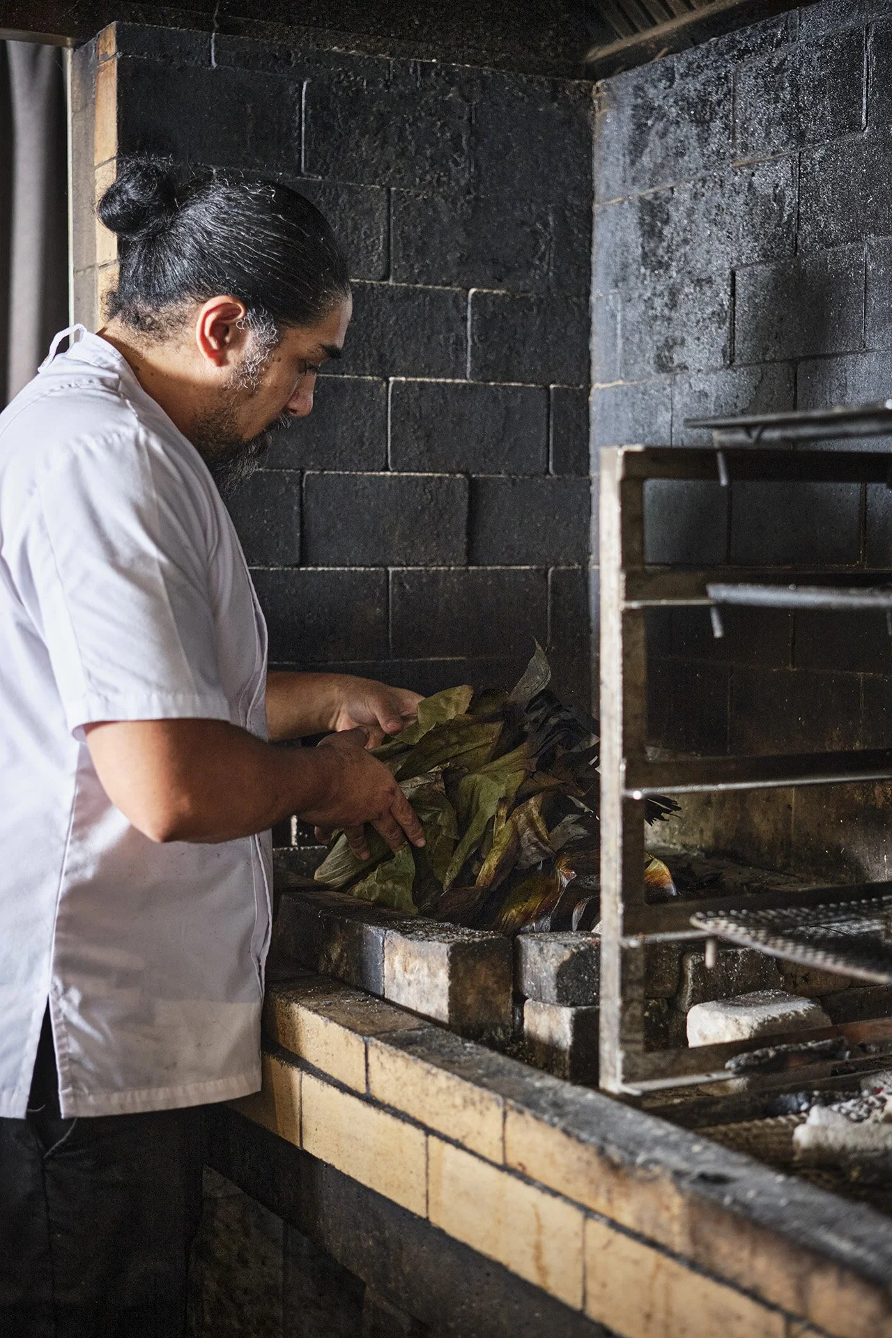A chef wearing a white apron stands in front of a brick oven, preparing food wrapped in banana leaves.
