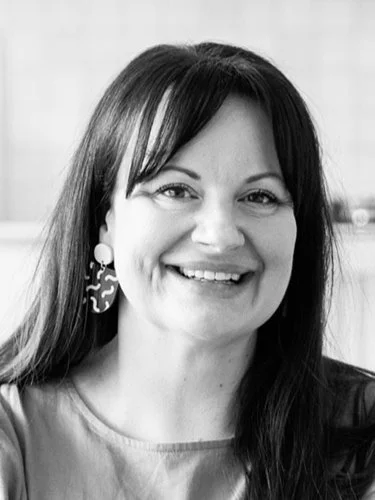 Black and white portrait of a woman with long dark hair, smiling, wearing earrings and a light-colored top.