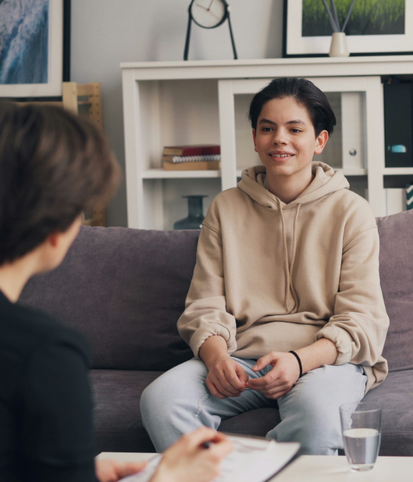 A young woman in a beige hoodie sitting on a gray sofa during a therapy session, engaging with a therapist who is taking notes, with a glass of water on the table.