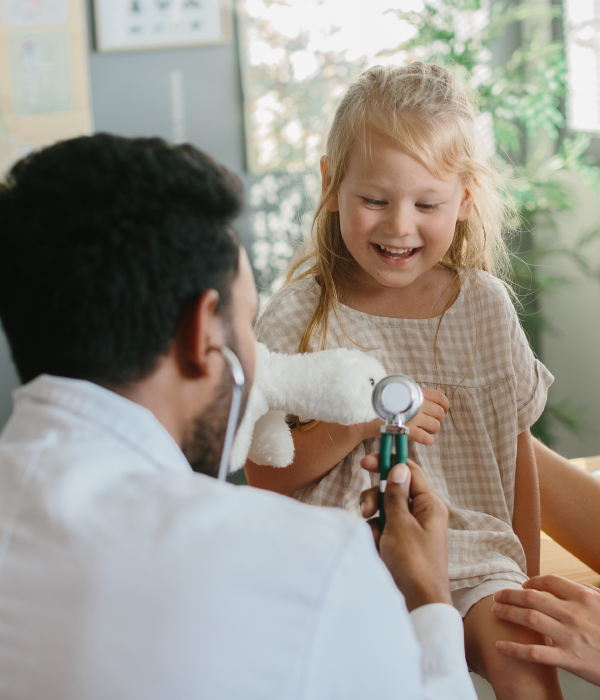 A young girl visiting the doctor and smiling while the doctor uses a stethoscope to listen to her chest.