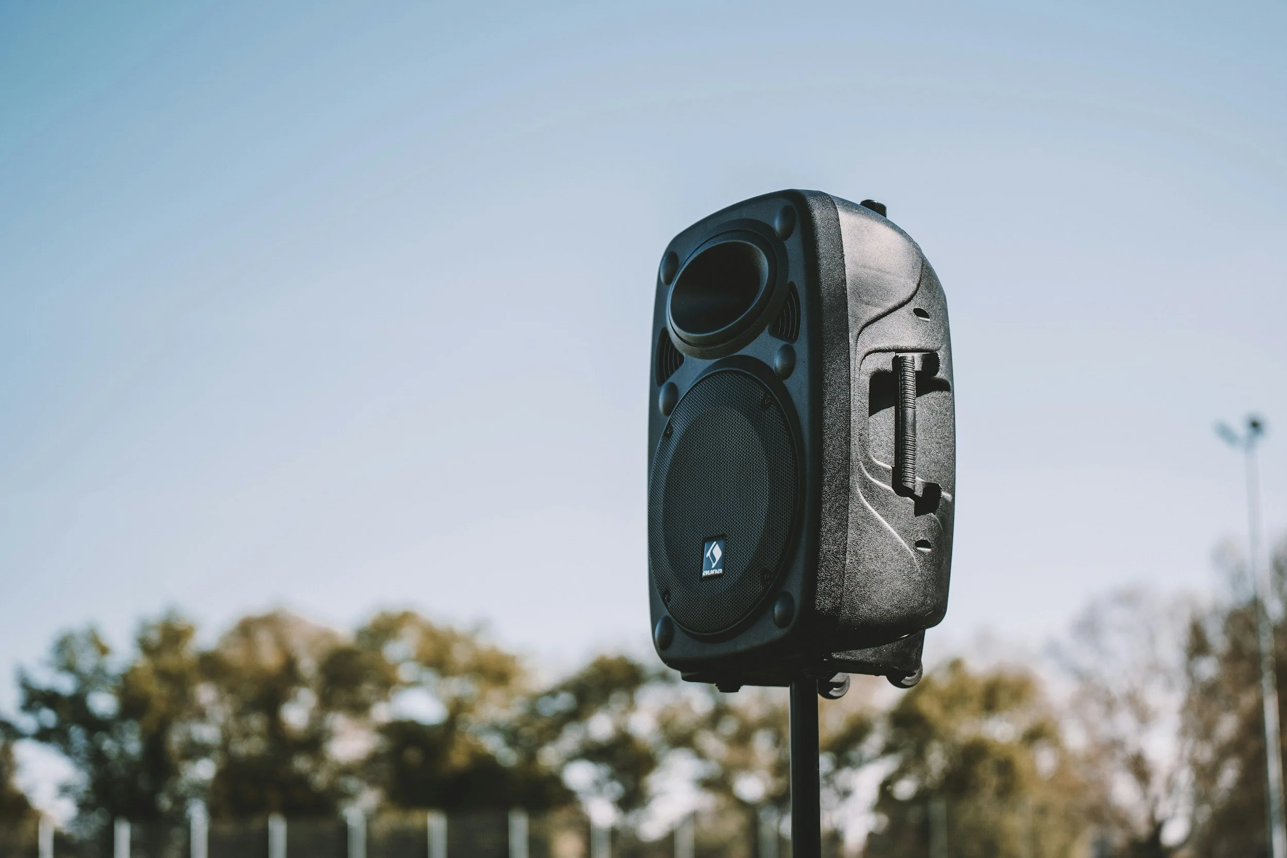 A black loudspeaker mounted outdoors on a stand against a clear sky with trees in the background.