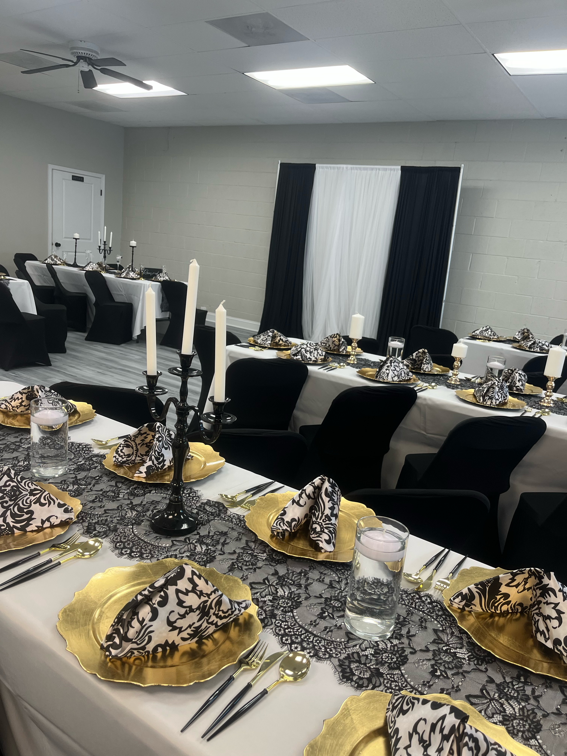 A long banquet table set for a formal event with black, white, and gold decorative elements. The table is covered with a gold and black patterned table runner and a black and white lace tablecloth underneath. Each place setting includes a fabric napk