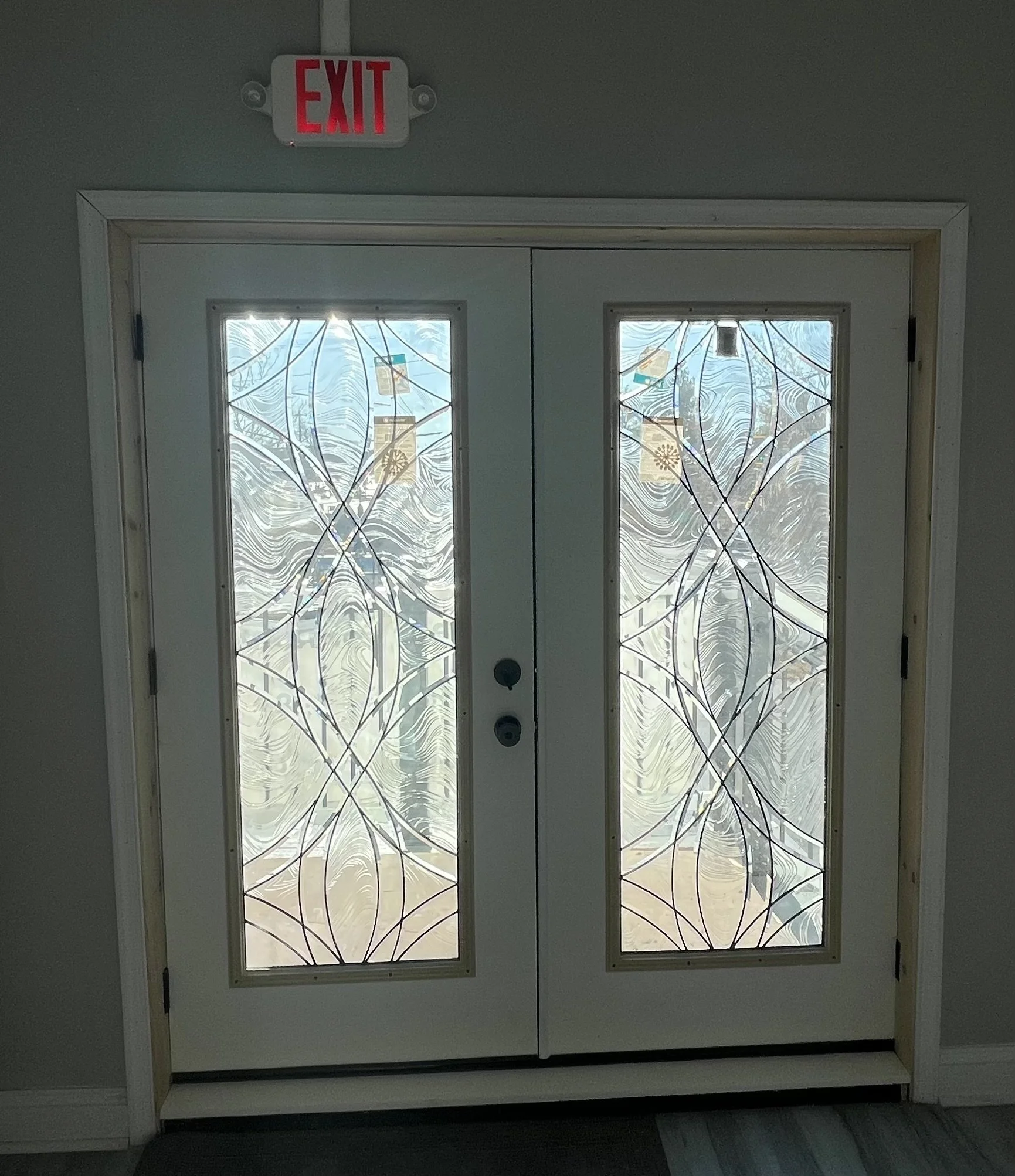 Double glass doors with decorative frosted glass design and a wooden frame, above which is a red exit sign. Outside the glass doors, a tree and some street scene are visible.