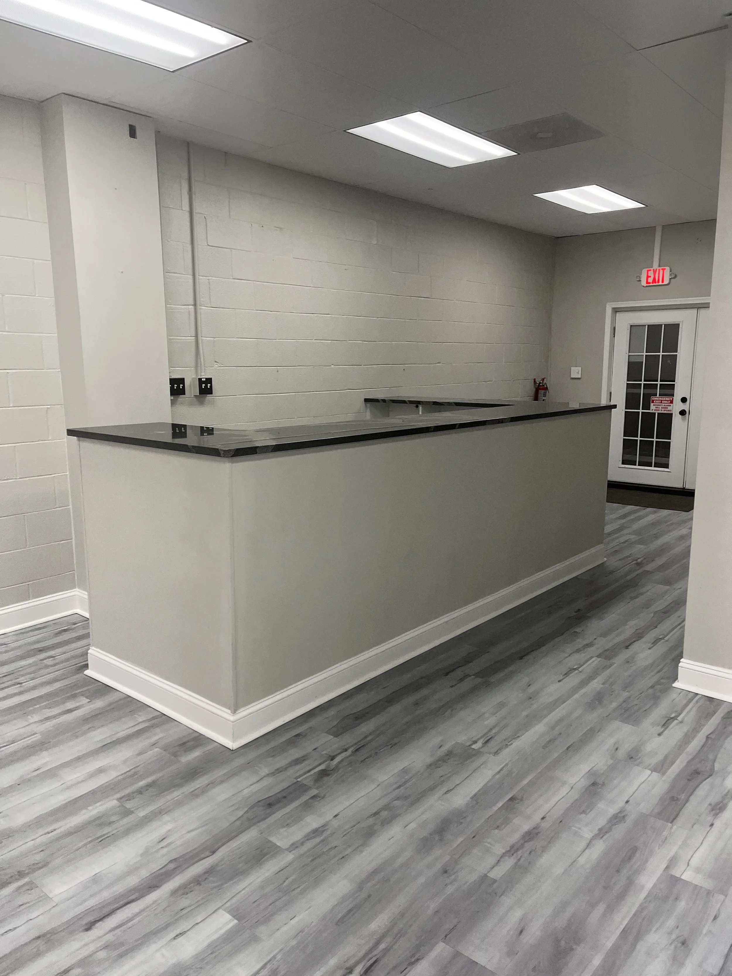 An empty indoor space with a counter, hardwood flooring, a white brick wall, ceiling lights, and an exit door with a red exit sign.