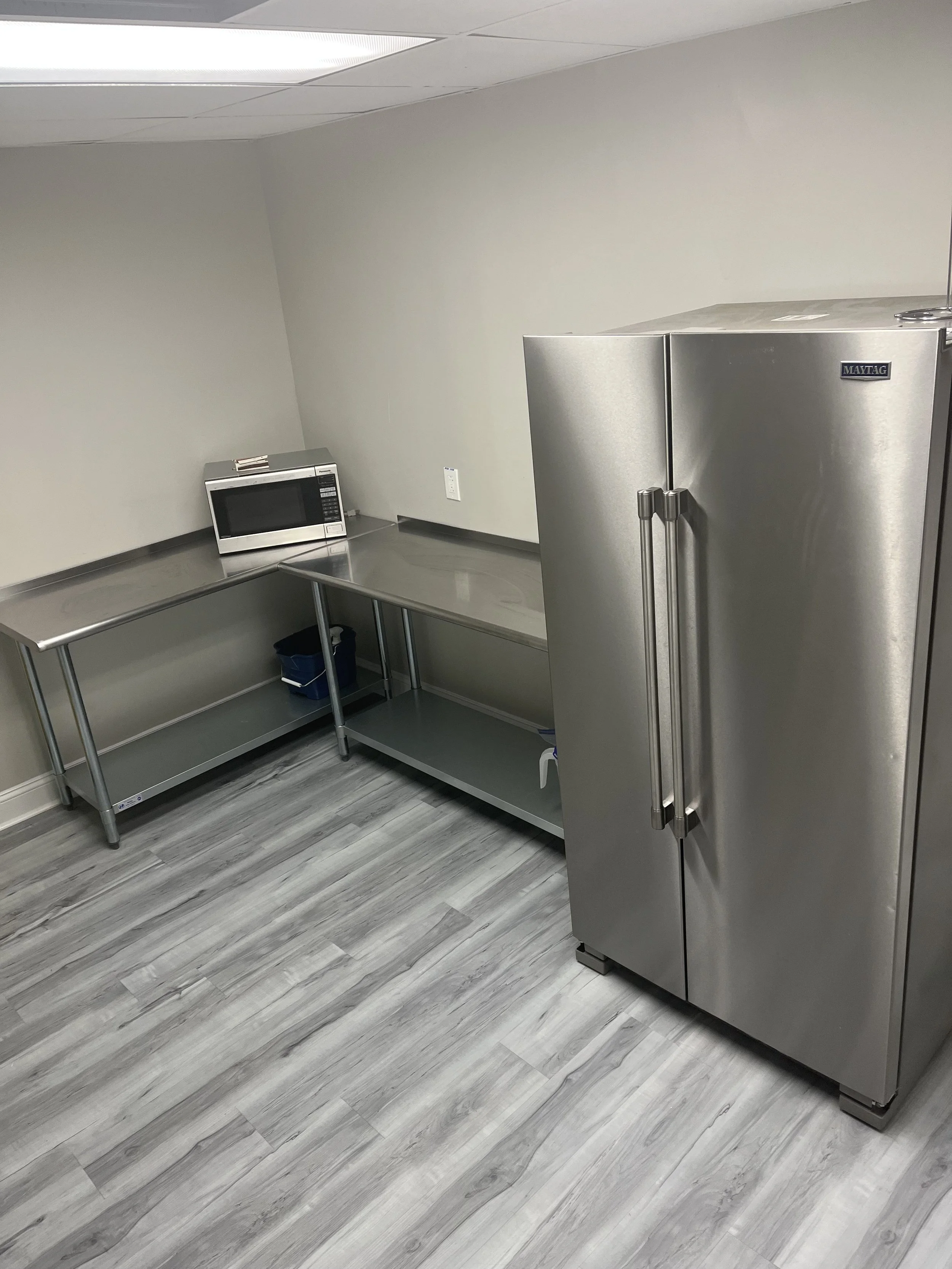 A kitchen or break room with a stainless steel refrigerator, a stainless steel table, a microwave, and wood-look flooring.
