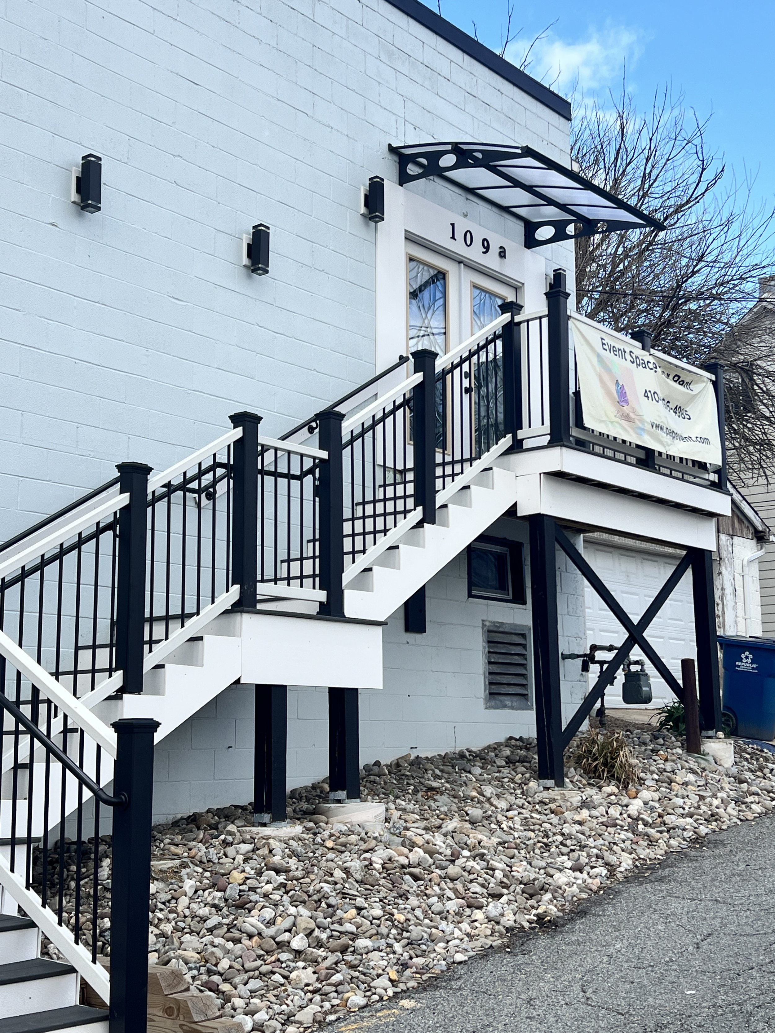 A two-story building with a white exterior wall and a black and white metal staircase leading to a door. The door has the number 109a above it. There are black wall-mounted lights on the wall, a canopy above the door, and a banner that reads 'Event S
