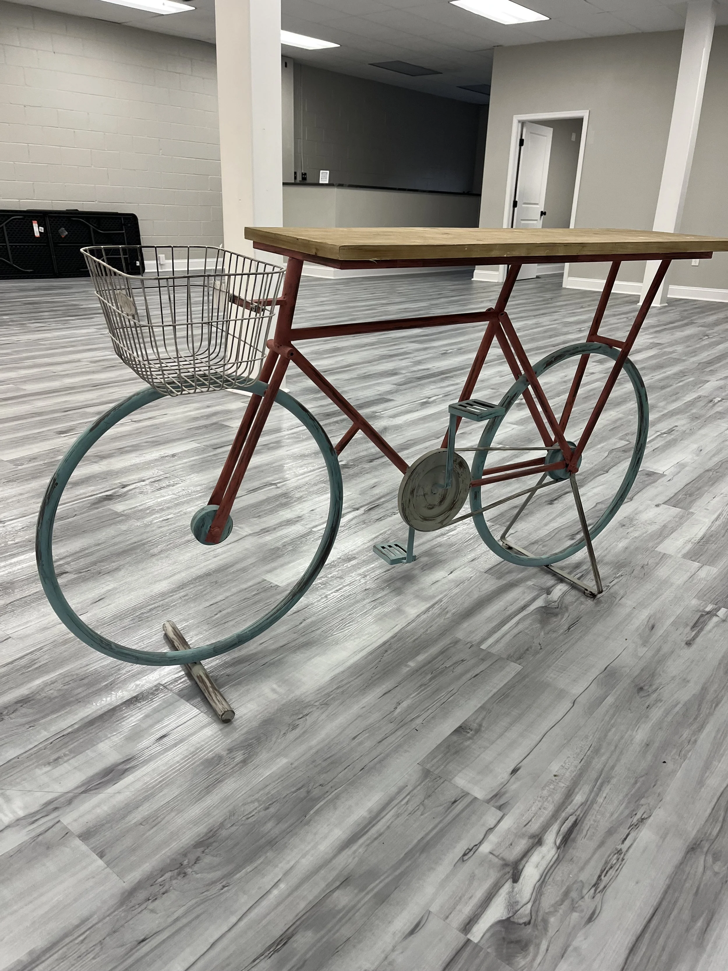 Empty indoor space with a wooden cart featuring a metal basket and large wheels, set on a gray wood-patterned floor, with white and gray walls and ceiling lights.
