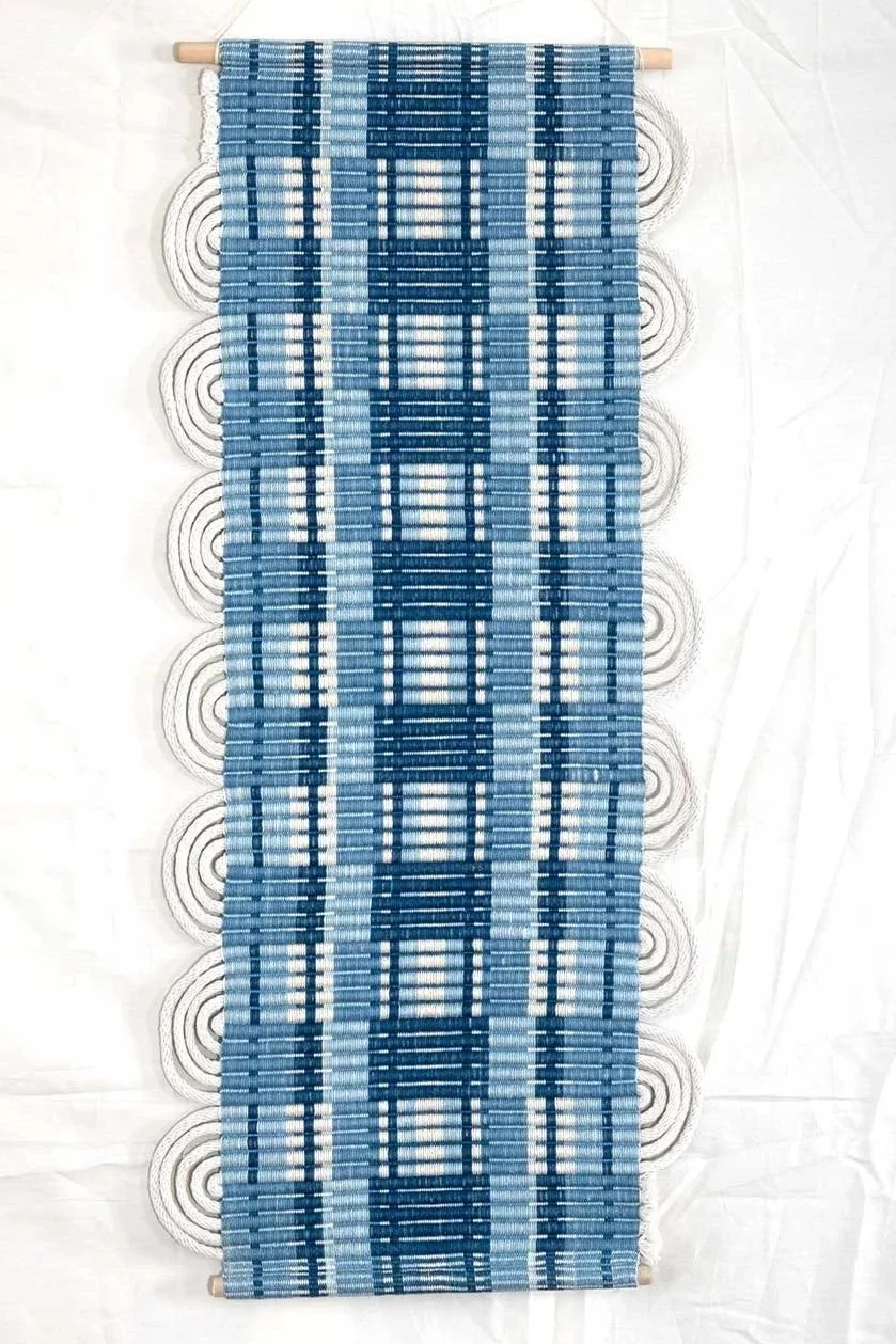 handwoven rep wall hanging, indigo dyed cotton yarns in create a series of stripes and rectangles on the surface of the piece, white cotton clothesline as the weft creates half circles in the style of passementerie, the piece is hung on wooden dowels