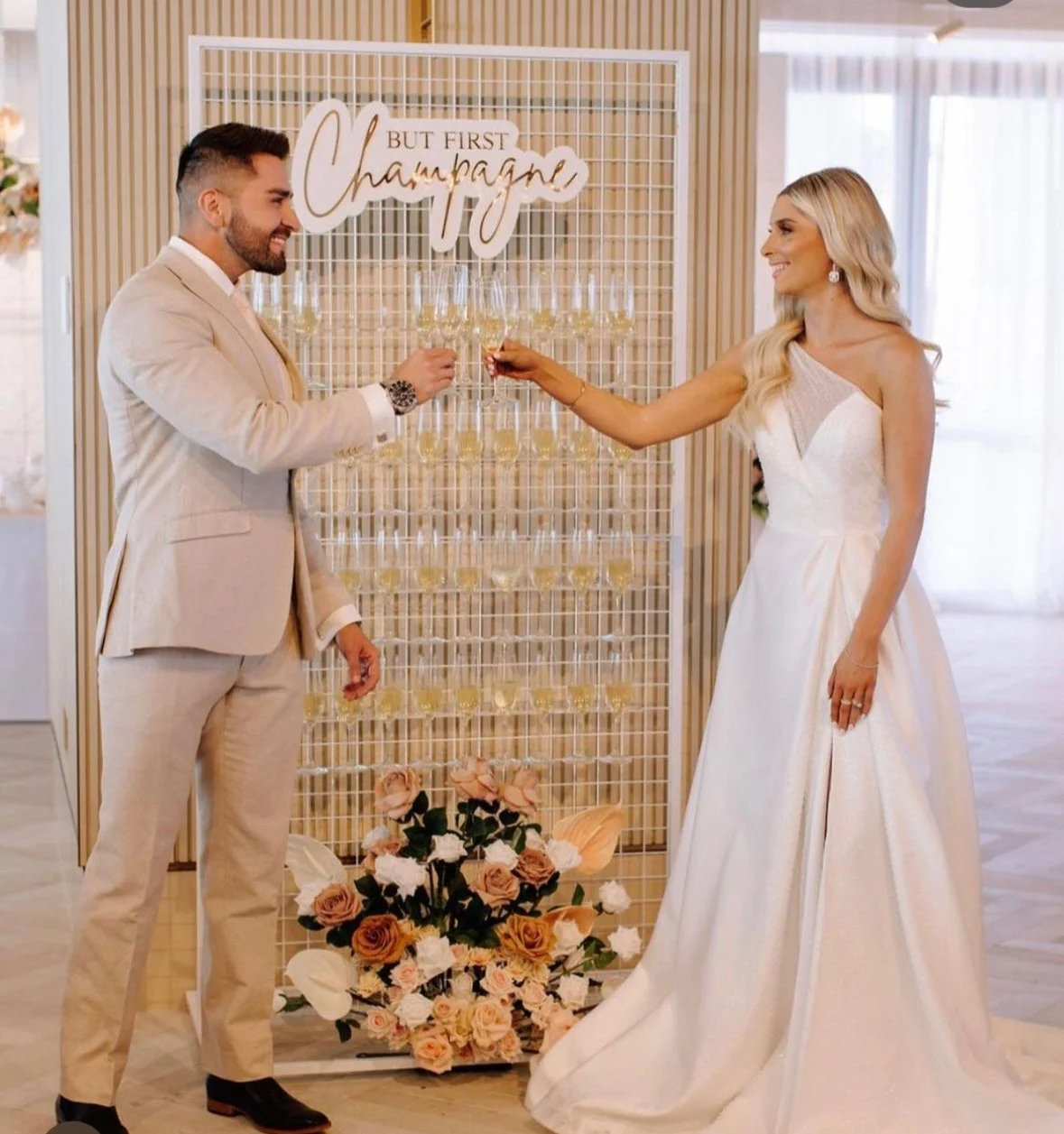 A bride and groom in formal attire clinking champagne glasses in front of a backdrop decorated with flowers and a sign that reads, 'But First, Champagne.' The bride is in a white dress and the groom is in a beige suit.