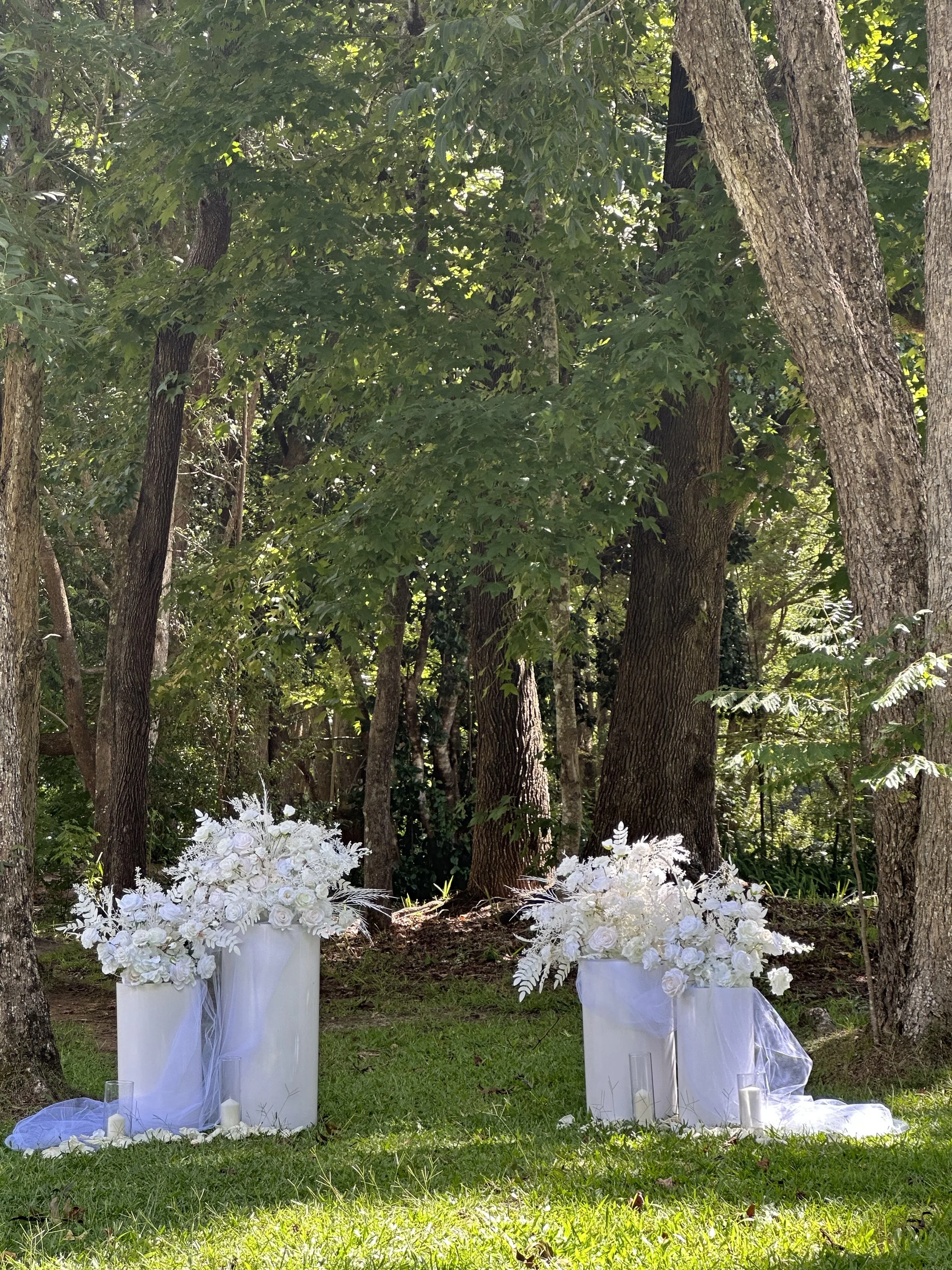 White floral arrangements on cylindrical vases draped with tulle, set in a forested area with trees and grass.