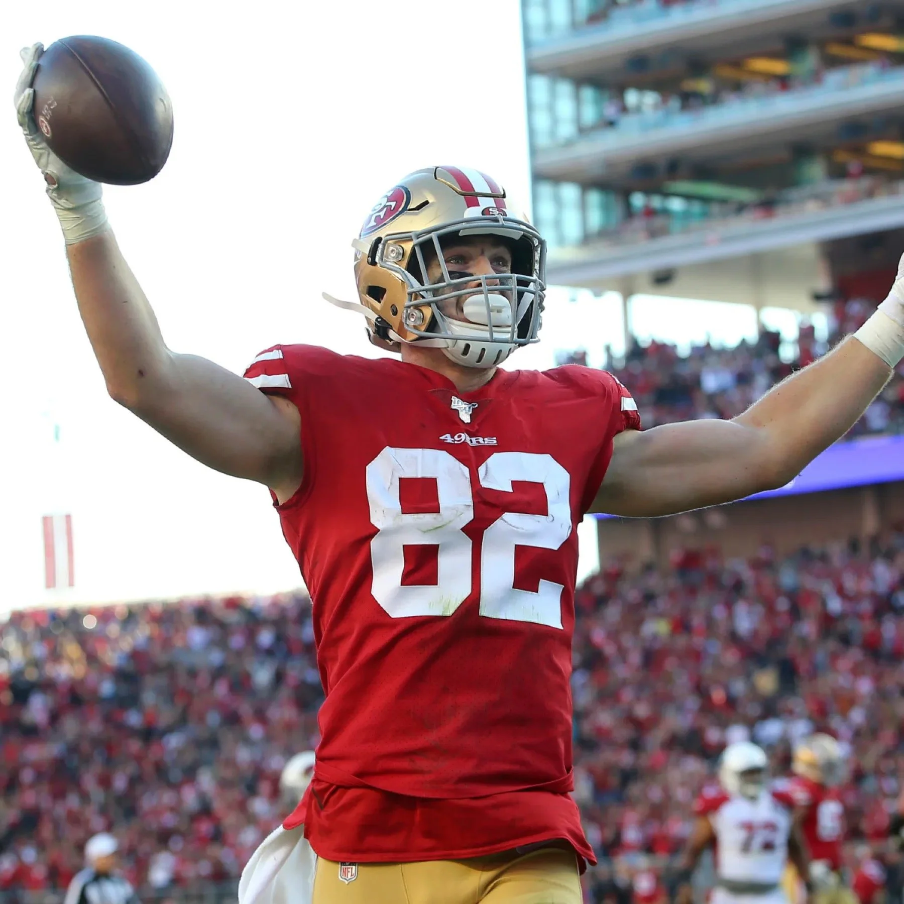 San Francisco 49ers football player wearing a red number 82 jersey and gold helmet, raising a football during a game in a packed stadium.