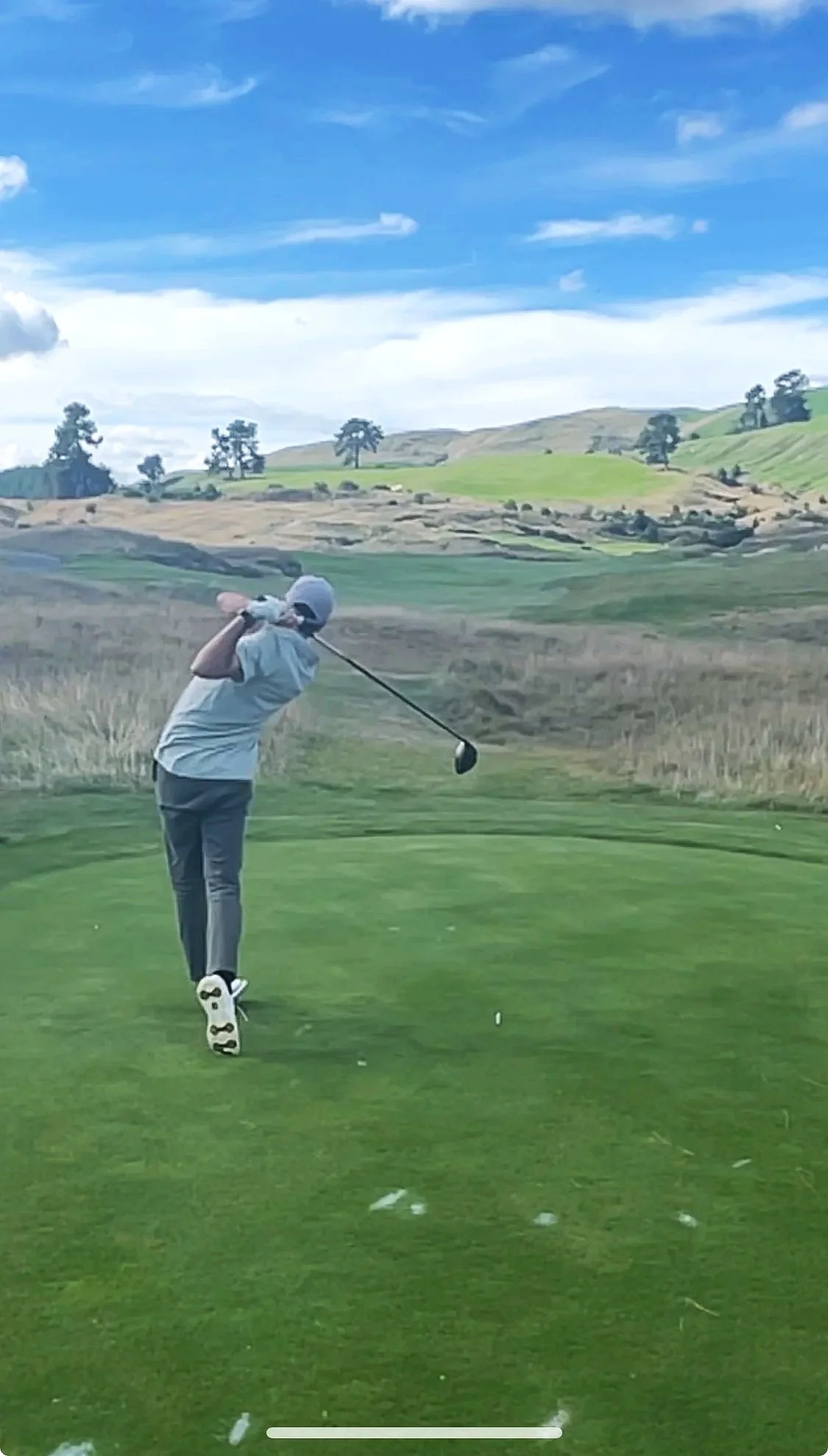 A person dressed in a light gray shirt, dark gray pants, and a white cap taking a swing with a golf club on a golf course with rolling hills and sparse trees under a partly cloudy sky.