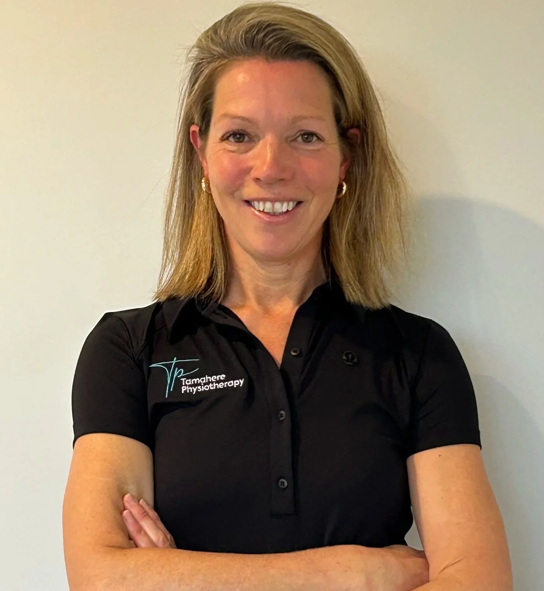 A woman with shoulder-length blonde hair wearing a black polo shirt with 'Tamahere Physiotherapy' logo, standing with arms crossed in front of a plain white wall.