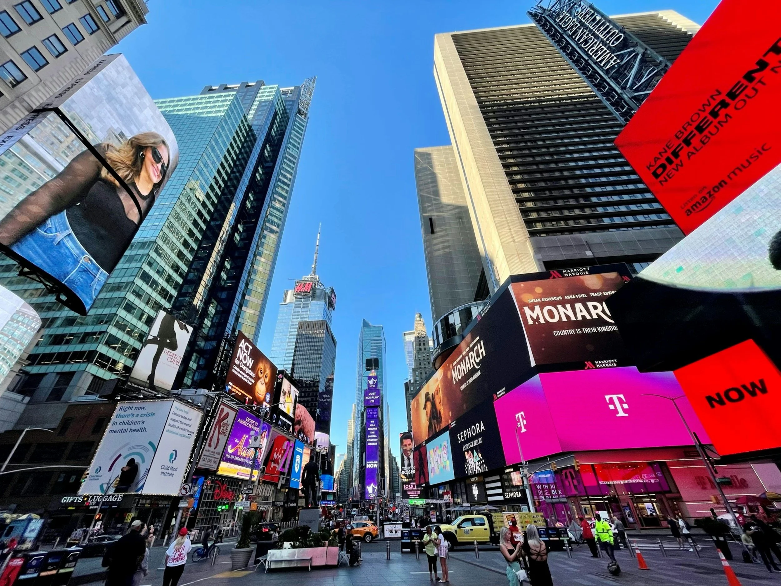 A bustling city square with tall buildings, bright billboards, and advertising screens, possibly Times Square in New York City, with people walking and vehicles on the street.