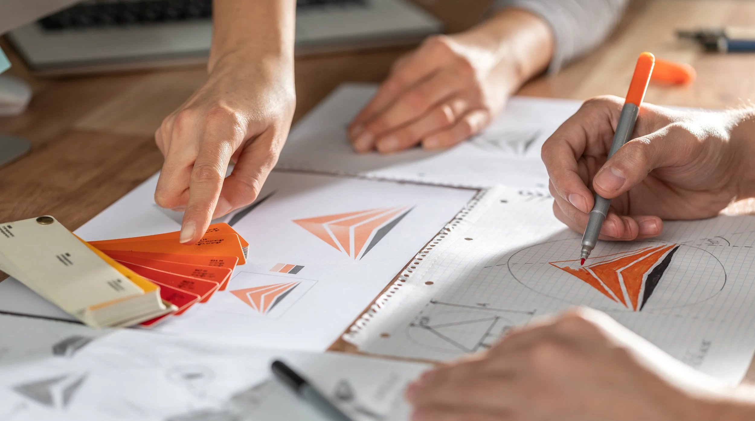 Designers working on logo concepts for a brand, reviewing color samples and sketches of a geometric logo on graph paper.