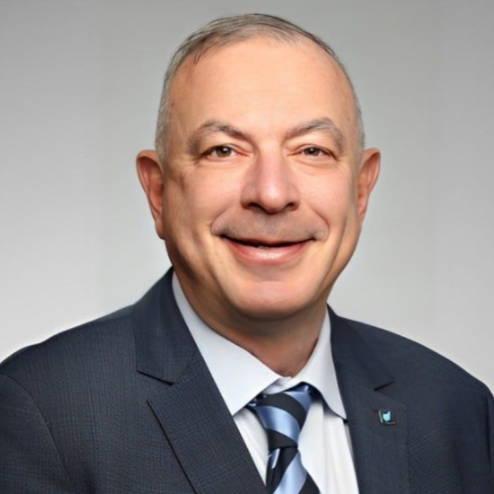 A professional headshot of a middle-aged man with short gray hair, wearing a dark suit, white shirt, and a striped tie, smiling at the camera against a plain light background.