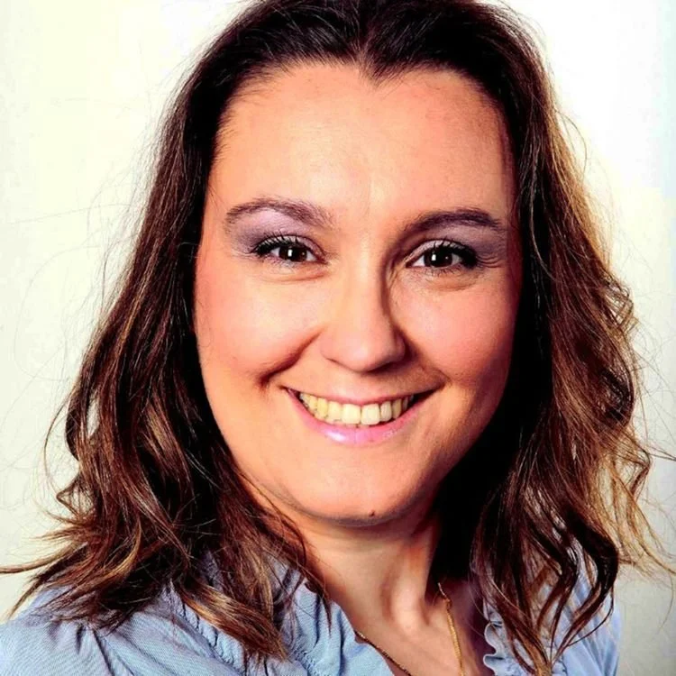 Smiling woman with shoulder-length brown wavy hair, wearing a light blue shirt and a gold necklace.