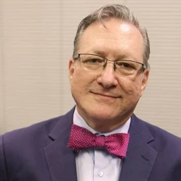 A middle-aged man wearing glasses, a dark blue suit, a white shirt, and a pink bow tie, smiling at the camera.
