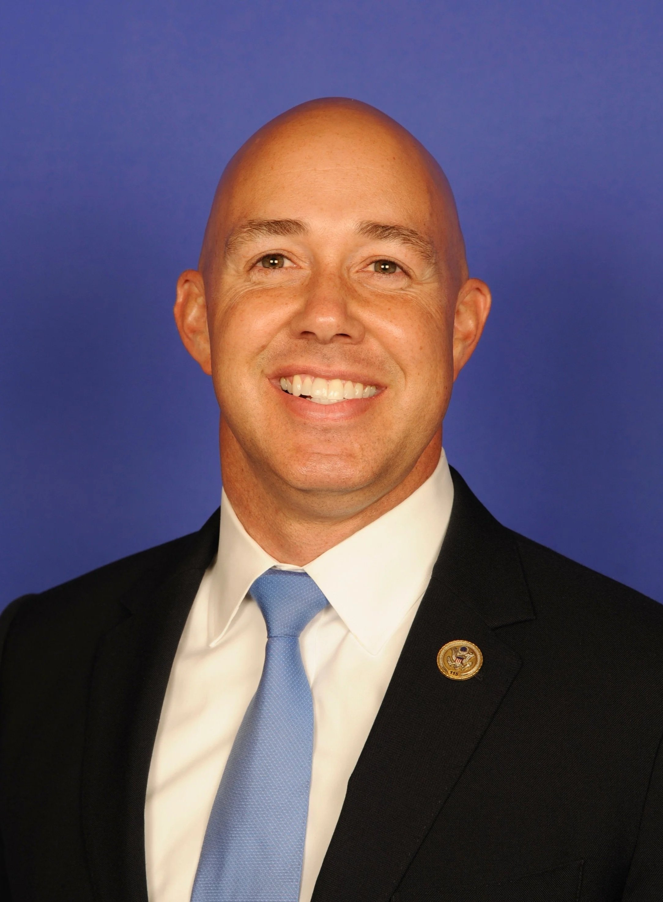 A smiling bald man in a black suit, white shirt, and light blue tie, with a U.S. congressional pin on his lapel, against a solid blue background.