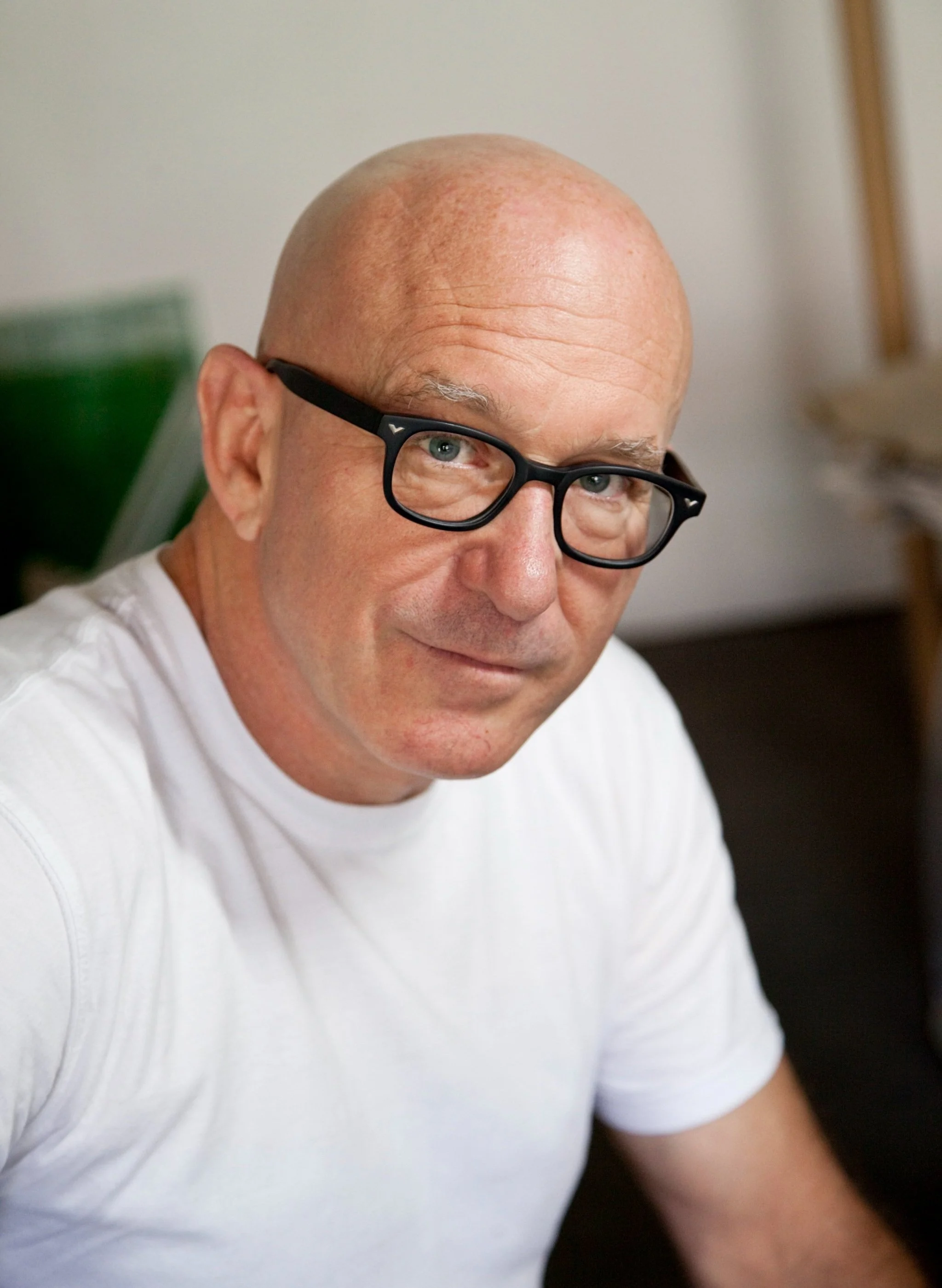 A middle-aged man with glasses and a shaved head, wearing a white T-shirt, looking at the camera with a slight smile in an indoor setting.
