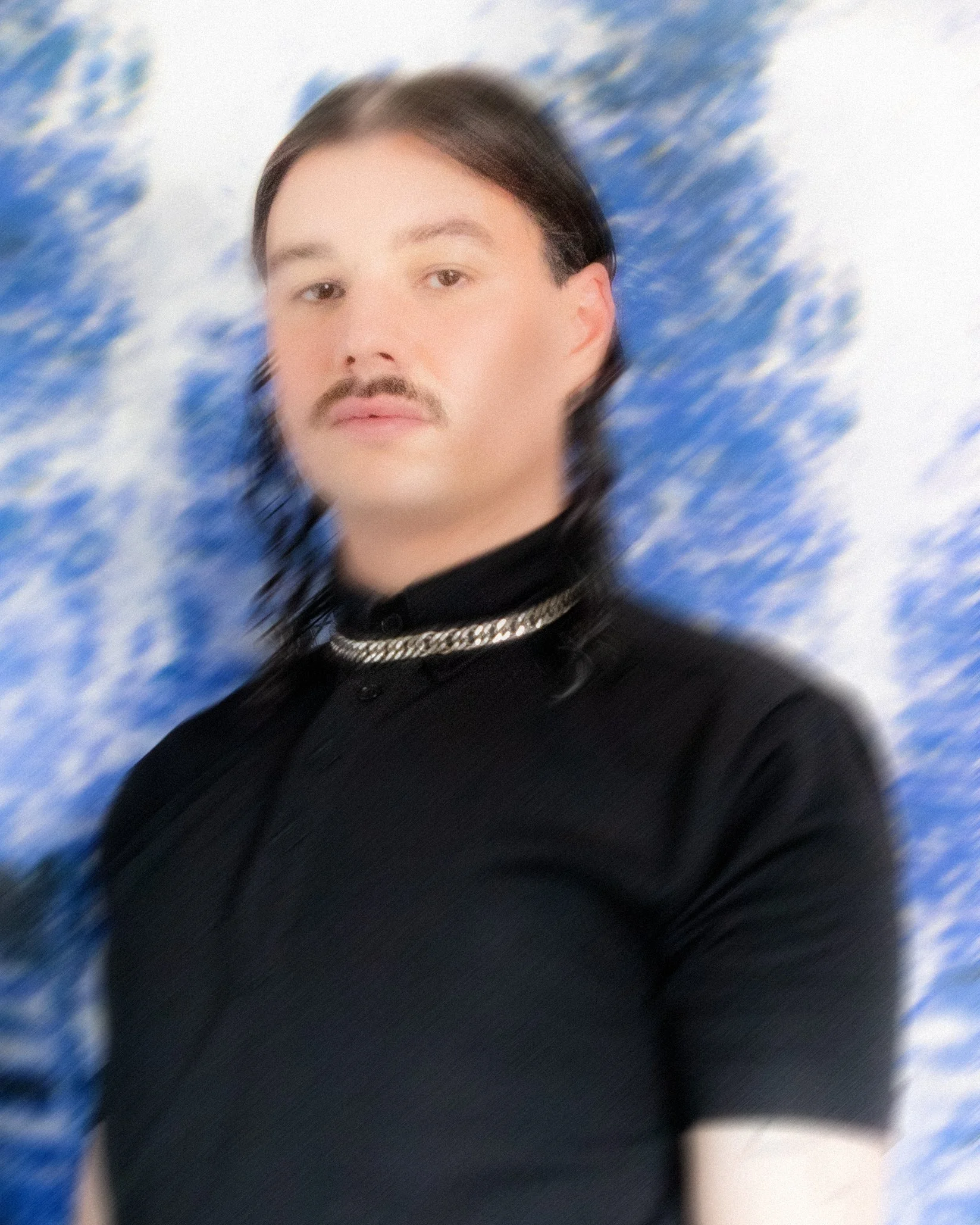 J. Mannherz mit dunklen, langen, lockigen Haaren, einem Oberlippenbart und einem schwarzen Oberteil, vor einem blauen Himmel mit Wolken.