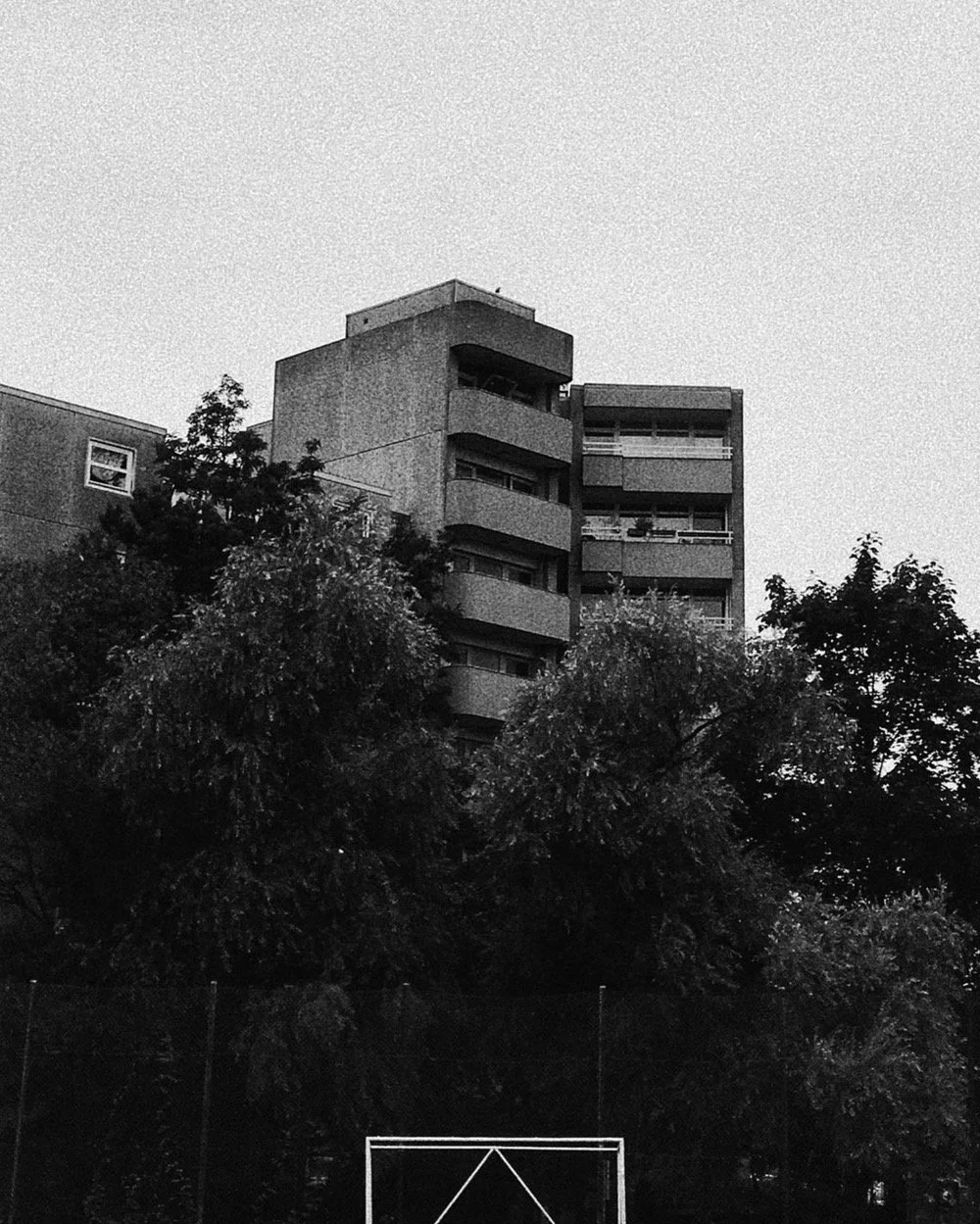 Ein mehrstöckiges Gebäude mit Balkon, im Vordergrund Bäume und ein Torzaun, schwarz-weiße Aufnahme.
