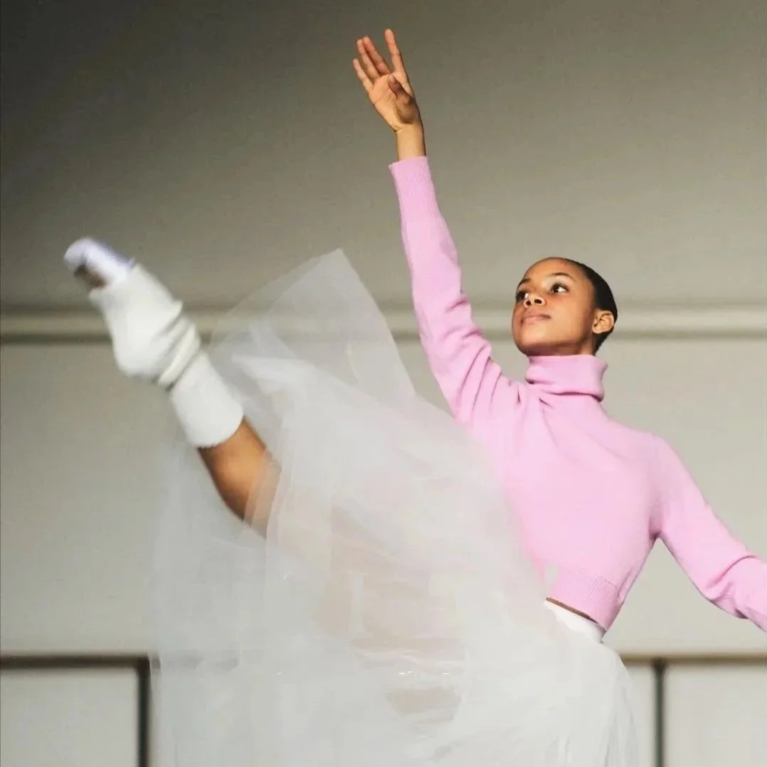 A young ballet dancer practicing in a studio, wearing a pink sweater, white tutu, and white socks, with one leg lifted and arms outstretched.