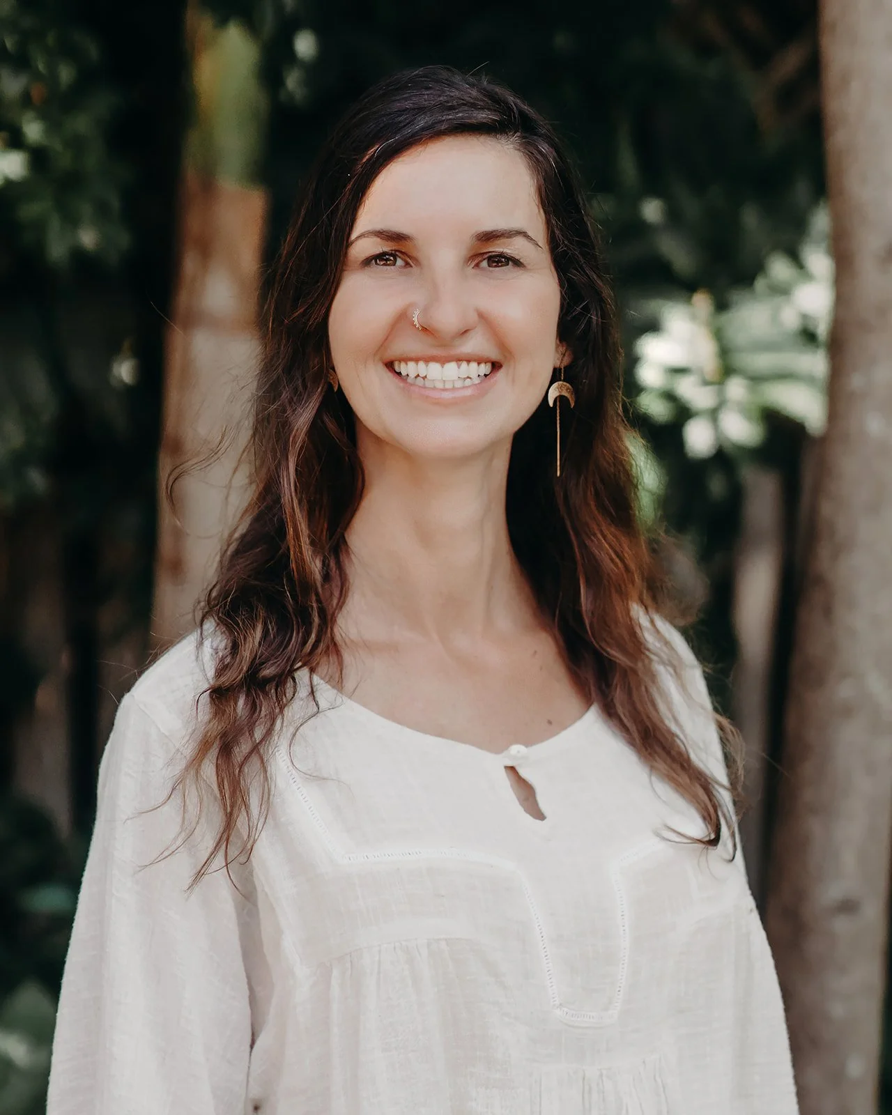 A woman with dark wavy hair smiling outdoors, wearing a cream-colored blouse and gold earrings, with trees in the background.