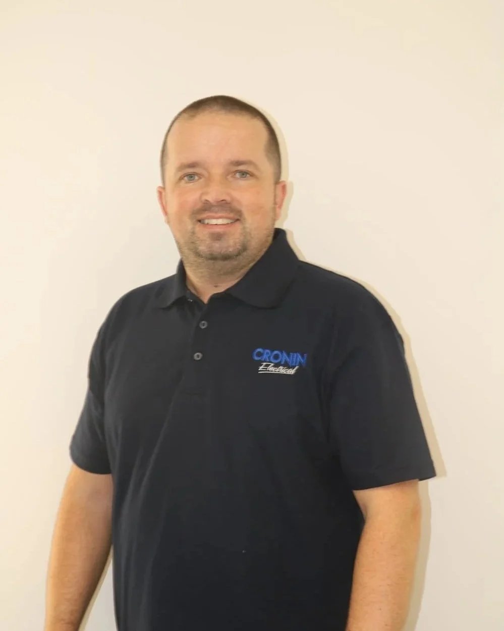 A man with short hair and a beard, wearing a black polo shirt with 'CROXN' embroidered on it, standing against a plain white wall, smiling.