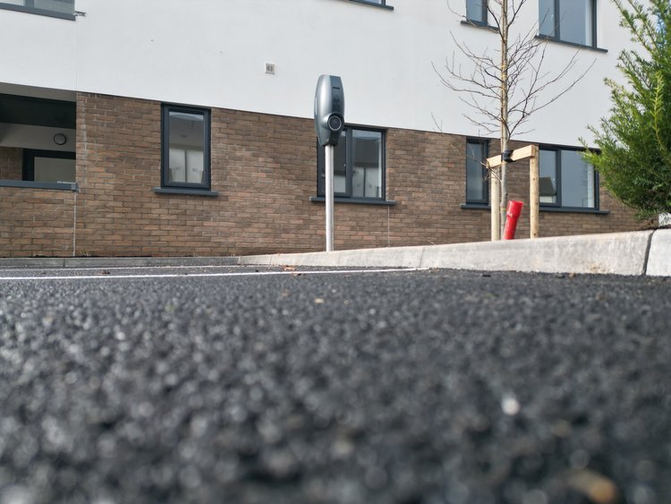 Low-angle view of a street with an electronic parking meter and a brick apartment building with windows, some trees, and a sidewalk.