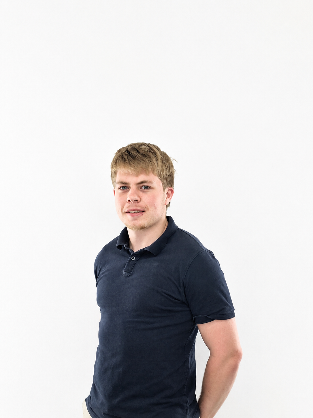 A young man with light brown hair and blue eyes wearing a dark blue polo shirt, standing against a plain white background.