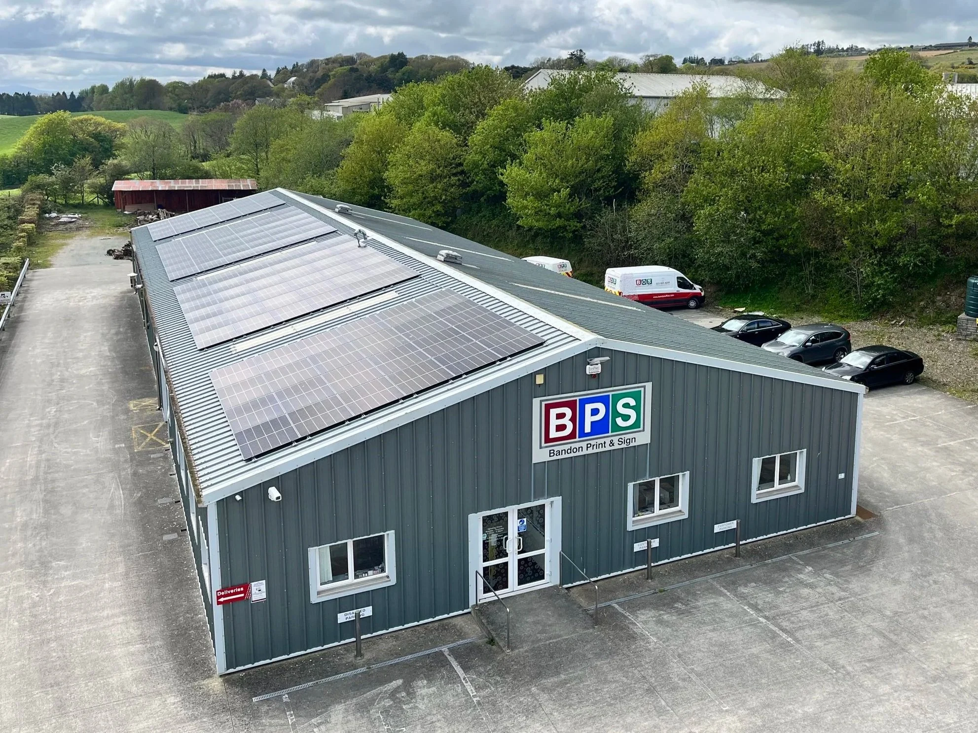An industrial Building with the sign Bandon Print & Sign has installed multiple solar panels on its roof. The parking lot in front of the building has several parked cars and vans, and surrounding trees and hills are visible in the background.