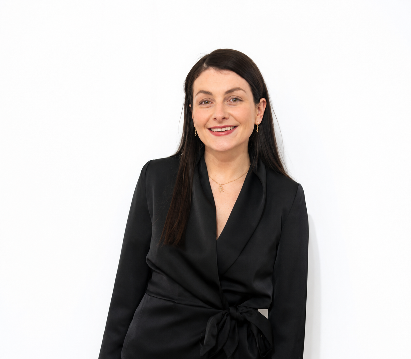 Portrait of a woman with long dark hair wearing a black satin blazer and jewelry, smiling against a white background.