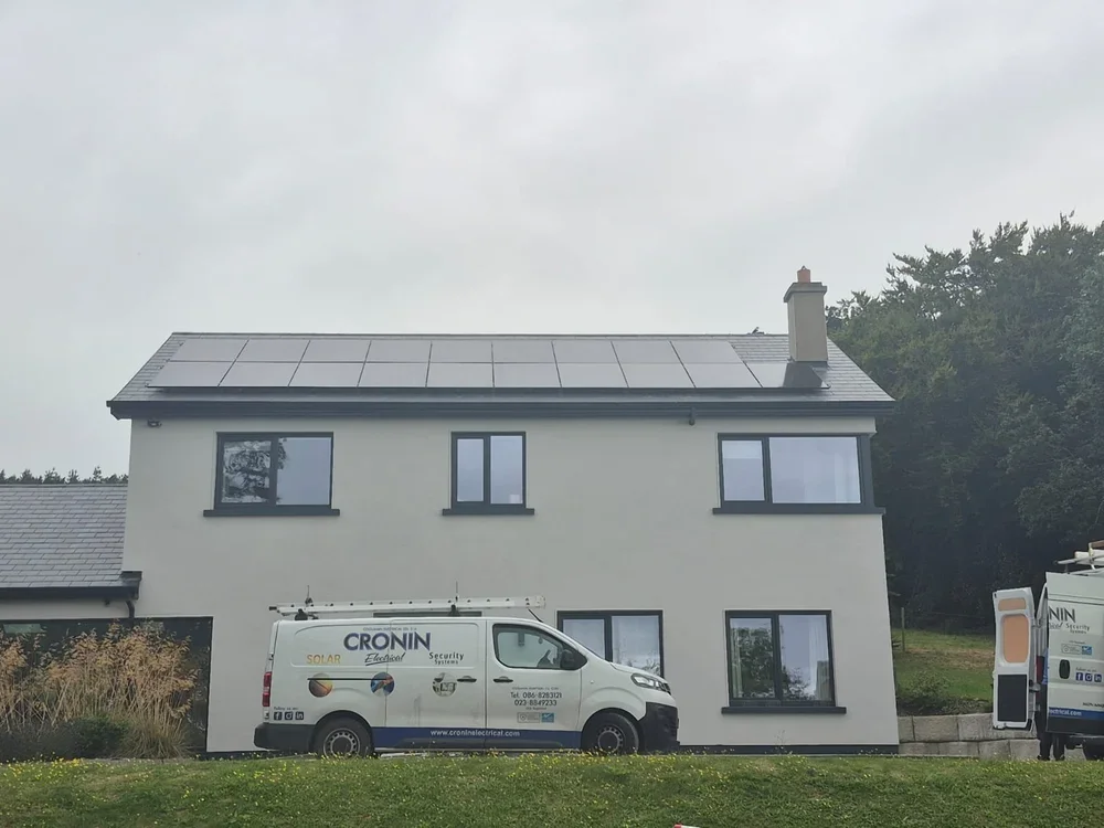 A white house with a dark roof and solar panels. There are five windows visible on the house. In front, there are two service vans, one of which has the words 'CRONIN Electrical' and 'Solar' written on it.