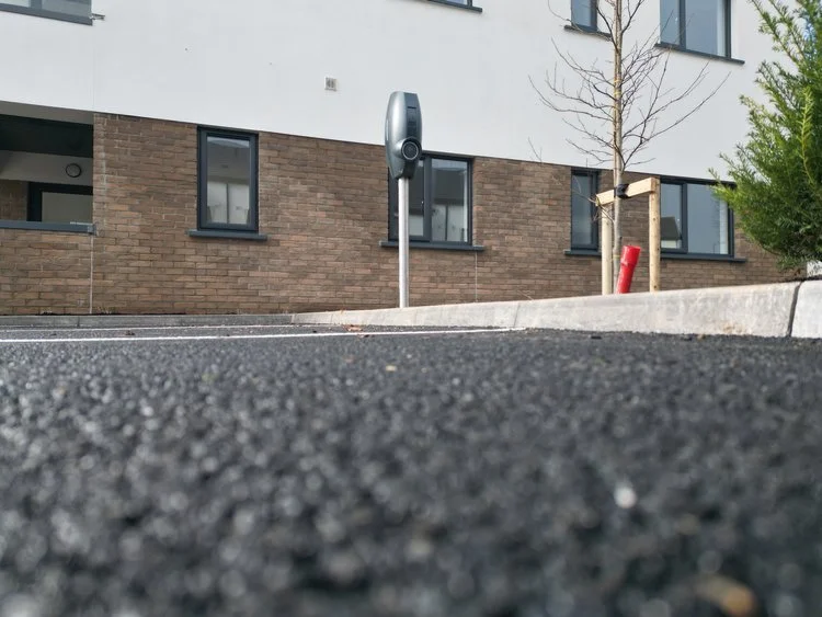A parking meter standing on the edge of a paved parking lot, with a modern building in the background, young trees, and green foliage visible.